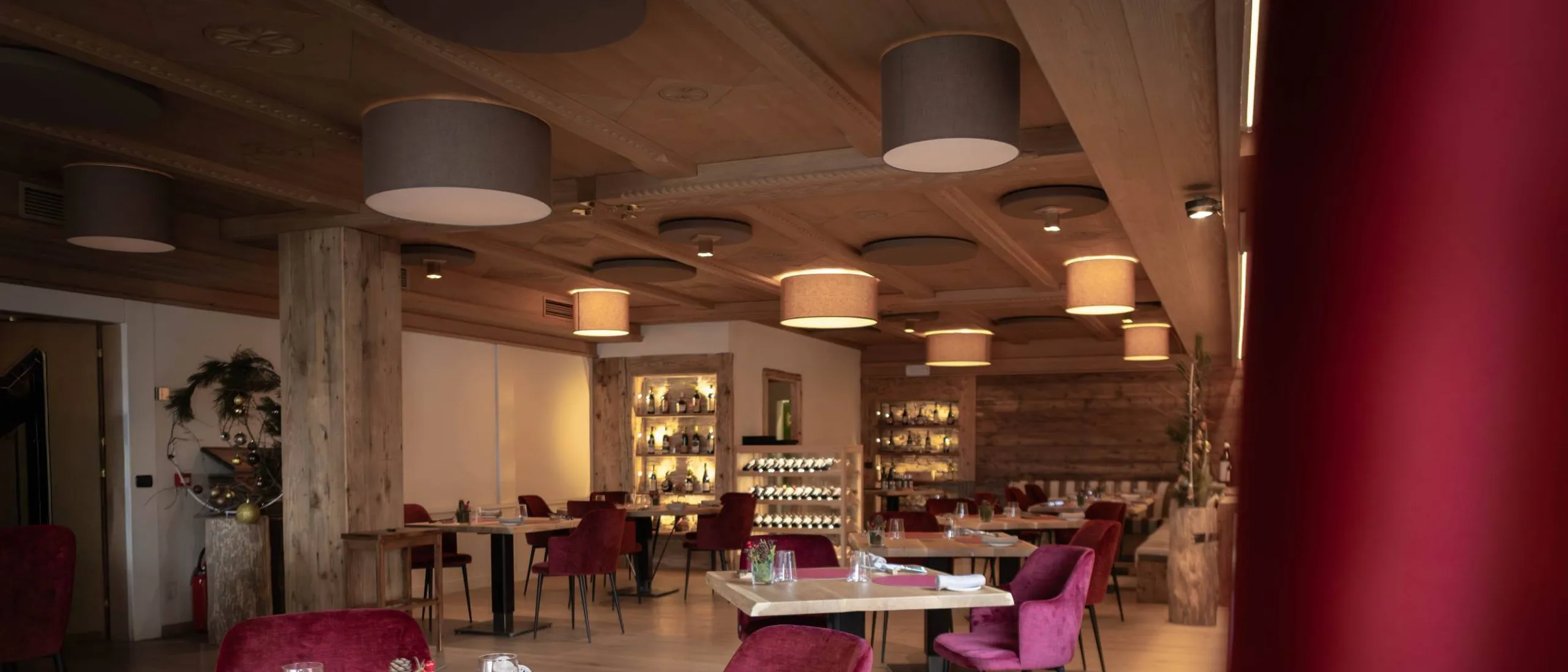 Sala ristorante moderna con soffitto in legno e grandi lampade circolari, tavoli rotondi in legno, sedie viola, piatti rosa e bicchieri; atmosfera calda e accogliente.