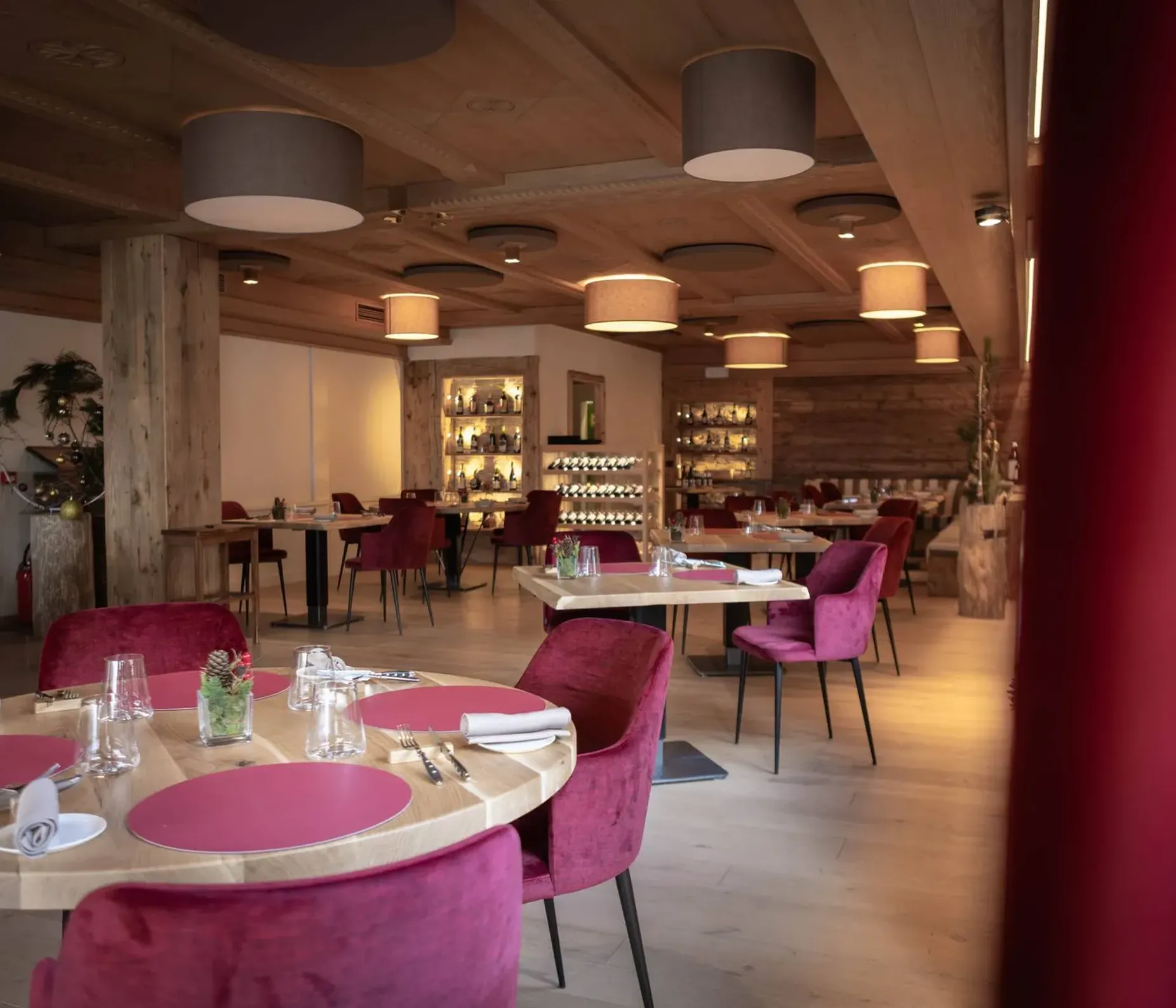 Sala ristorante moderna con soffitto in legno e grandi lampade circolari, tavoli rotondi in legno, sedie viola, piatti rosa e bicchieri; atmosfera calda e accogliente.
