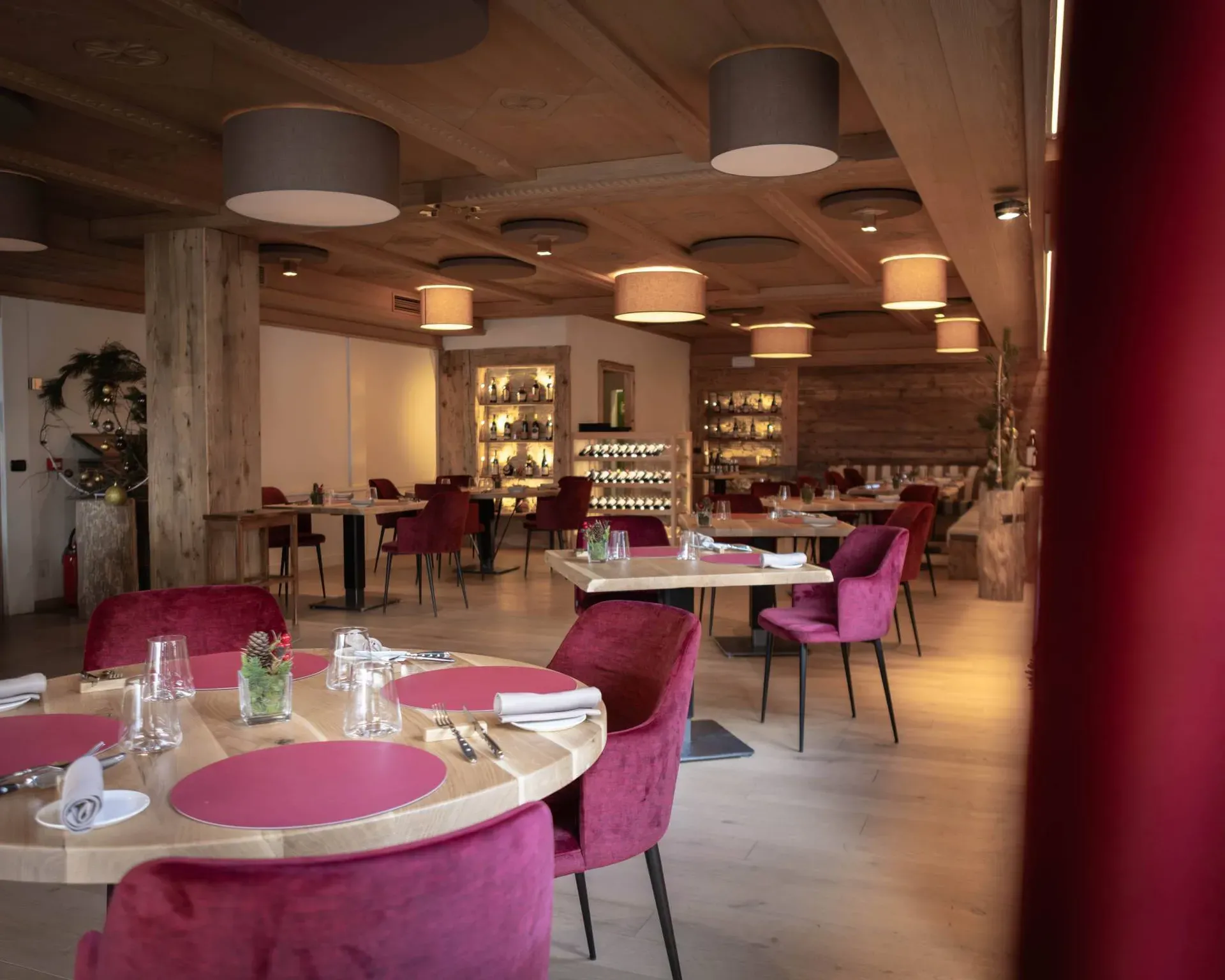 Sala ristorante moderna con soffitto in legno e grandi lampade circolari, tavoli rotondi in legno, sedie viola, piatti rosa e bicchieri; atmosfera calda e accogliente.