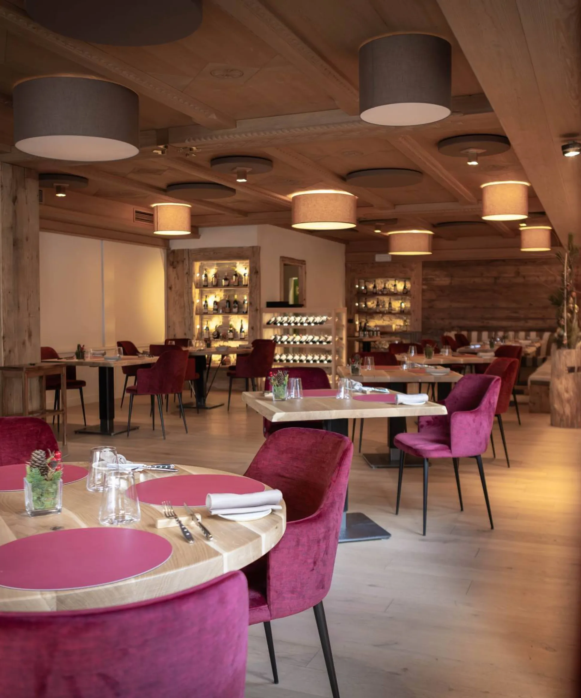 Sala ristorante moderna con soffitto in legno e grandi lampade circolari, tavoli rotondi in legno, sedie viola, piatti rosa e bicchieri; atmosfera calda e accogliente.