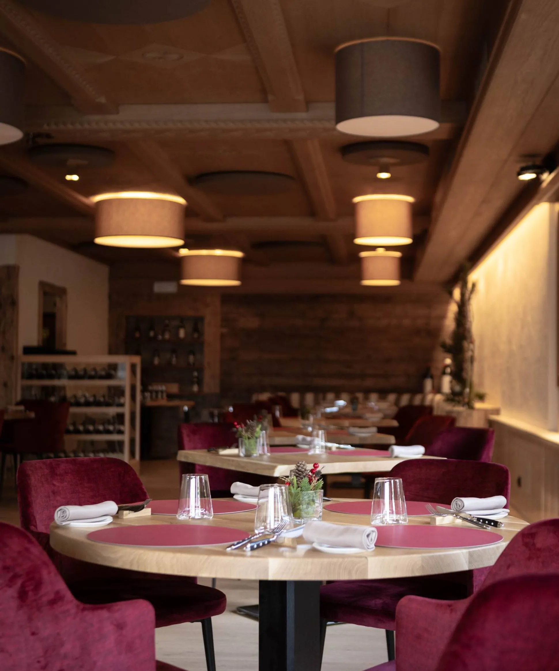 Interno di ristorante elegante con tavoli rotondi di legno, sedie velluto rosso, tovaglioli arrotolati e vasi di piante, luci pendenti calde.