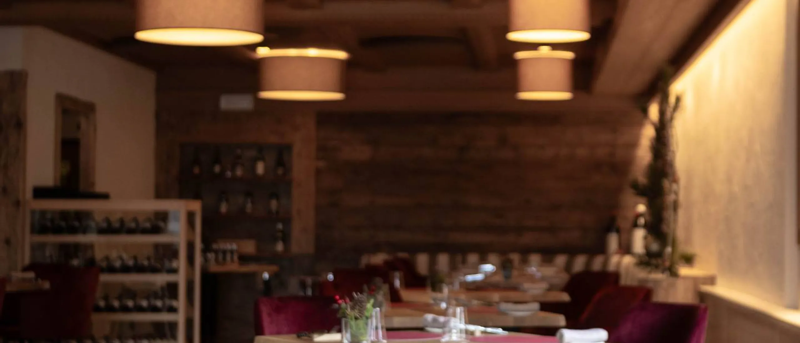 Interno di ristorante elegante con tavoli rotondi di legno, sedie velluto rosso, tovaglioli arrotolati e vasi di piante, luci pendenti calde.