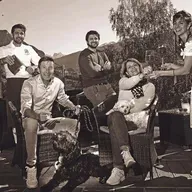 Gruppo di adulti su una terrazza all'aperto brindano e sorridono, con due cani tra sedie in rattan; montagne sullo sfondo in tonalità seppia.
