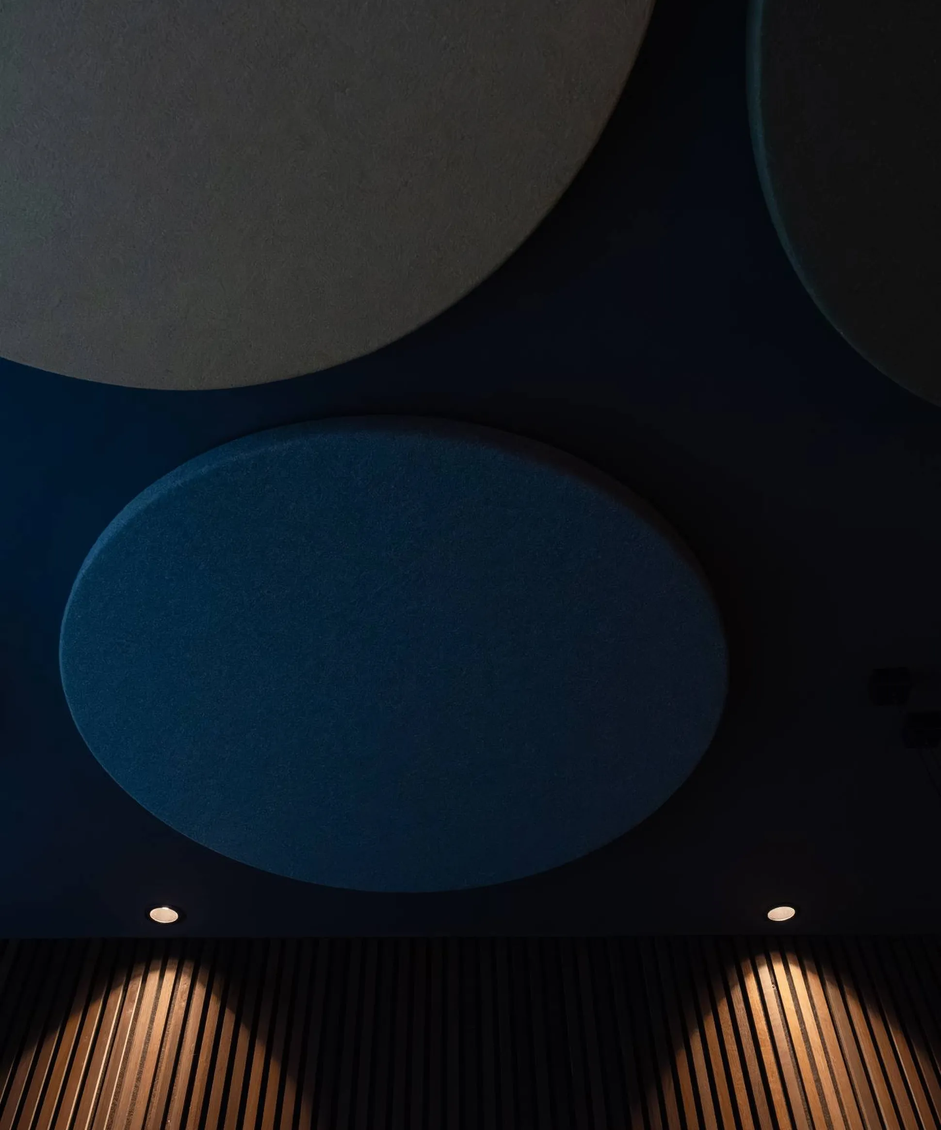 Interno con pannelli acustici circolari Silenzia sul soffitto, tonalità blu, faretti e pareti rivestite da listelli di legno.