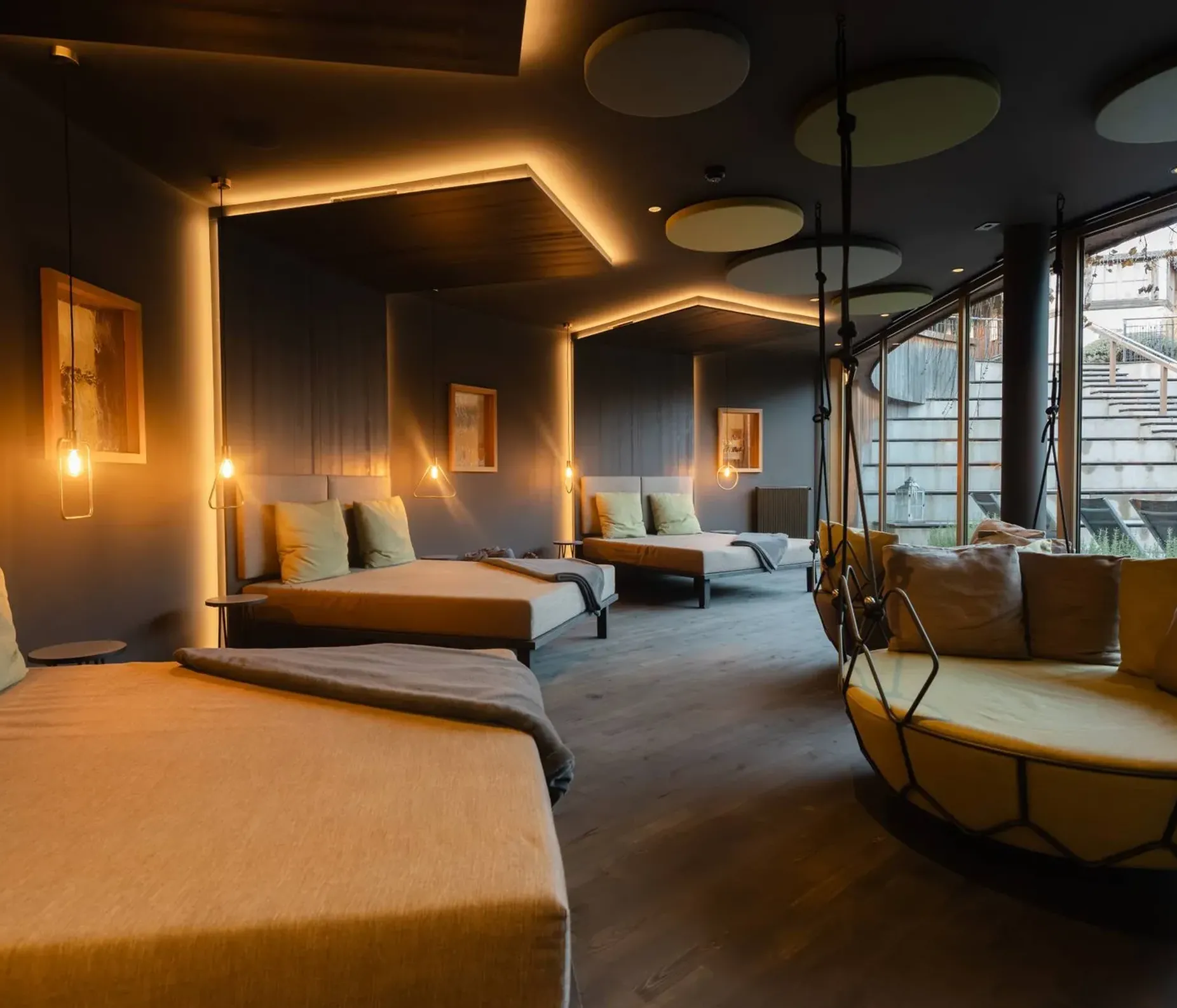 Interno moderno con letti lounge allineati lungo una parete blu, luci pendenti calde, soffitto con pannelli circolari, grande poltrona sospesa vicino alla vetrata.