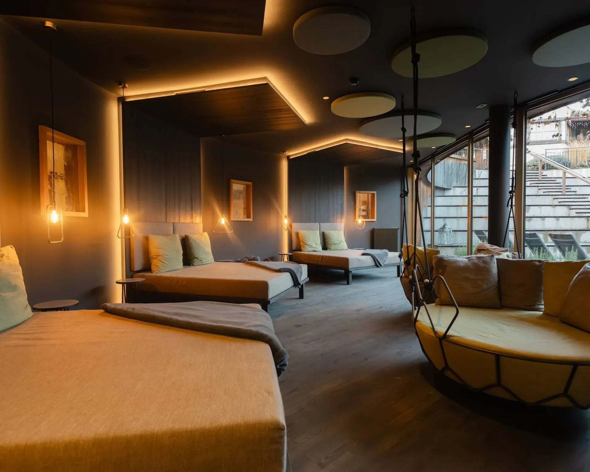 Interno moderno con letti lounge allineati lungo una parete blu, luci pendenti calde, soffitto con pannelli circolari, grande poltrona sospesa vicino alla vetrata.