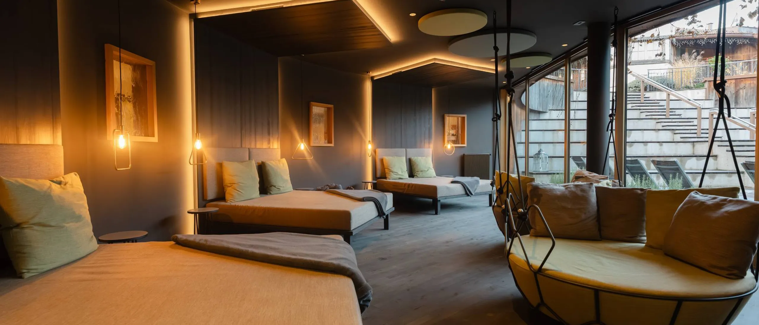 Interno moderno con letti lounge allineati lungo una parete blu, luci pendenti calde, soffitto con pannelli circolari, grande poltrona sospesa vicino alla vetrata.