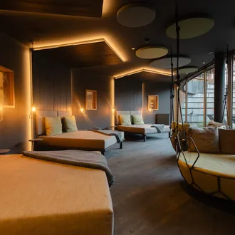 Interno moderno con letti lounge allineati lungo una parete blu, luci pendenti calde, soffitto con pannelli circolari, grande poltrona sospesa vicino alla vetrata.