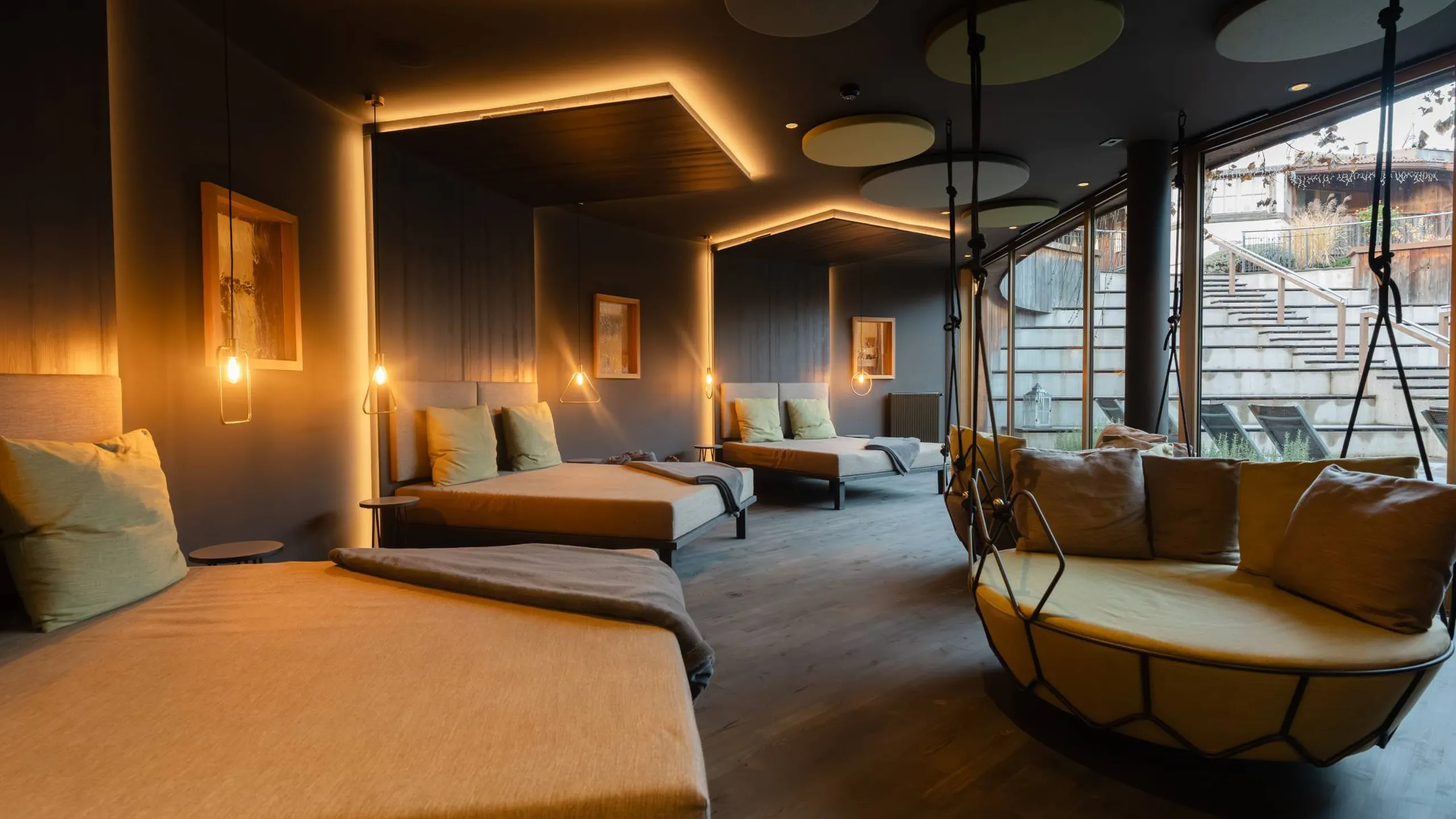 Interno moderno con letti lounge allineati lungo una parete blu, luci pendenti calde, soffitto con pannelli circolari, grande poltrona sospesa vicino alla vetrata.