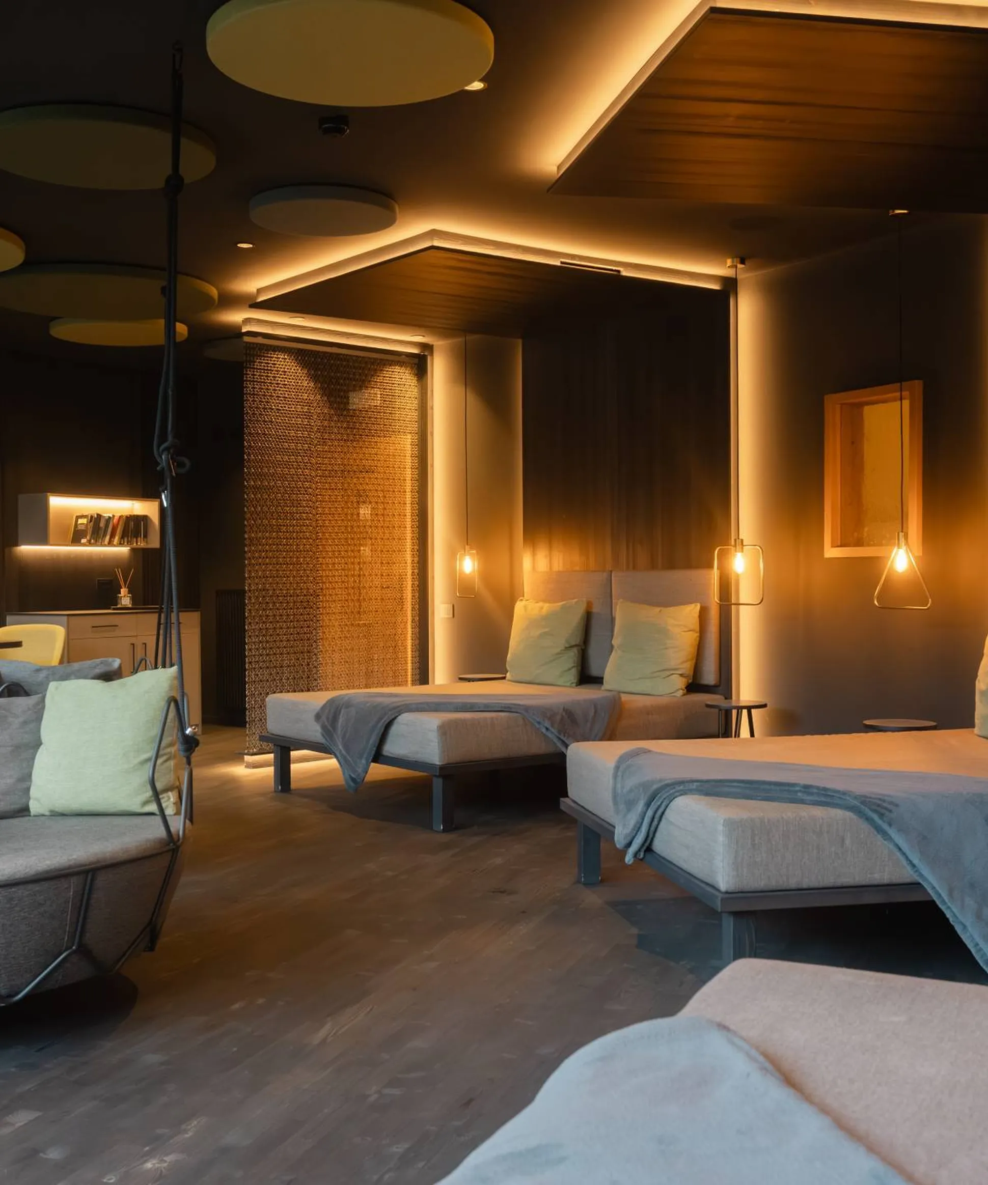 Lounge moderno: daybeds grigi con cuscini verde pallido, pouf sospeso rotondo e sedute laterali; luci calde su pareti scure con pannelli acustici.