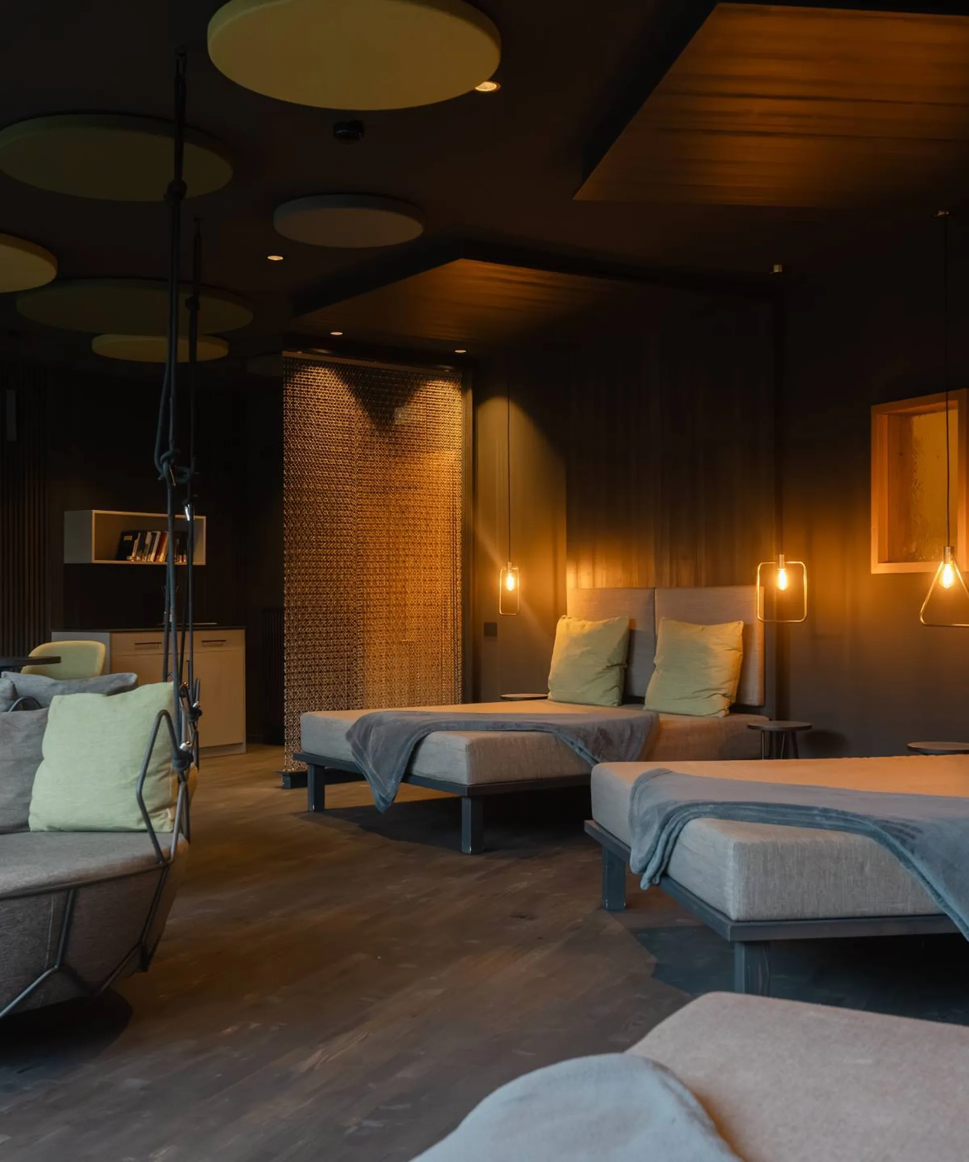 Lounge insonorizzato Silenzia: divani a piattaforma con cuscini verde chiaro, una poltrona sospesa rotonda, e luci ambrate su pareti di legno scuro.