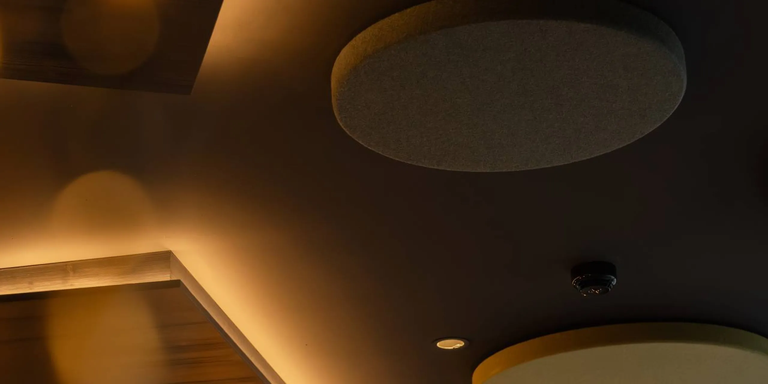 Interno con soffitto scuro, pannelli acustici circolari grigi e illuminazione calda che definisce i contorni; superfici in legno creano atmosfera accogliente.