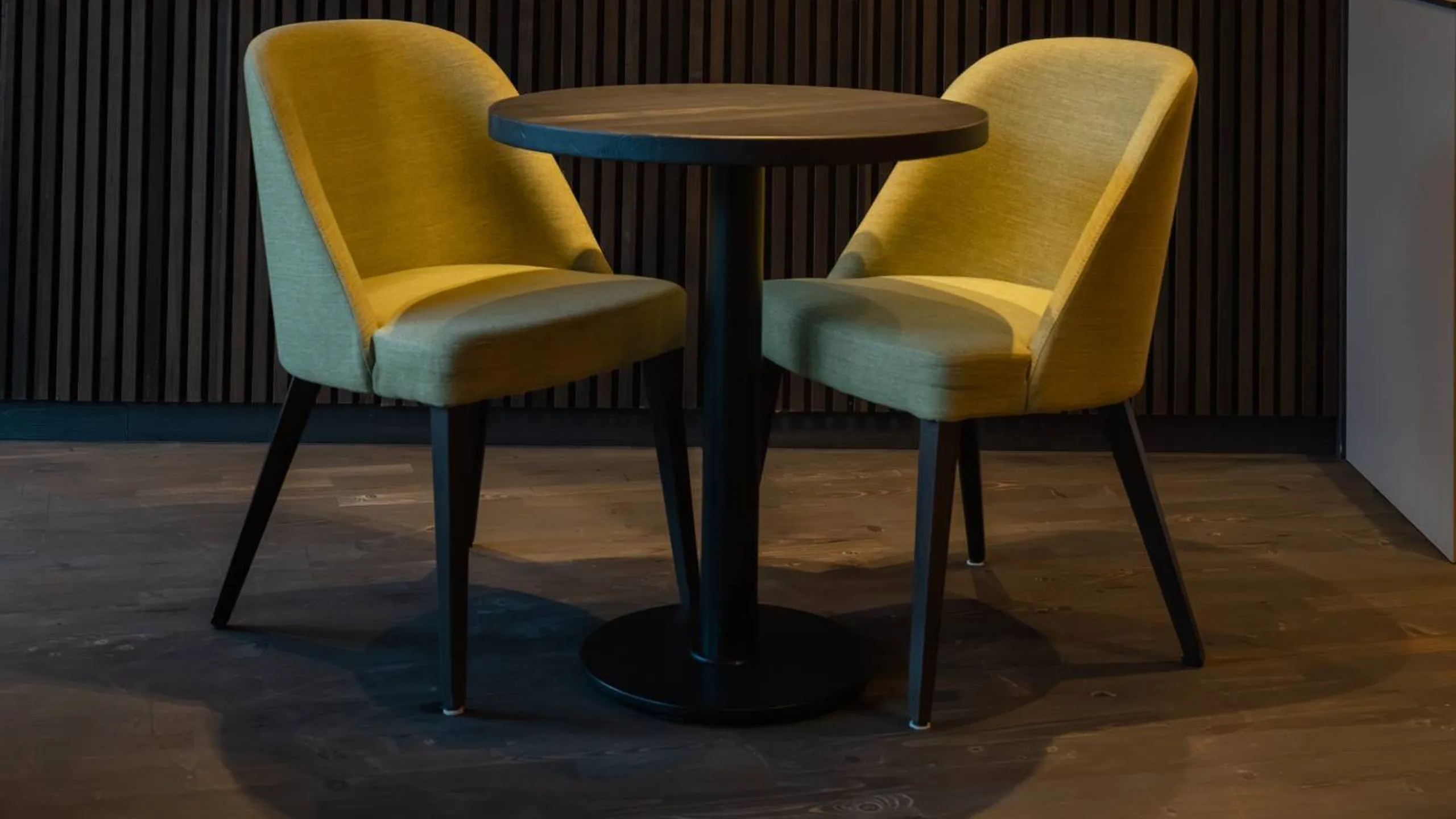 Due poltrone gialle disposte attorno a un tavolo rotondo nero, in una lounge con pannelli fonoassorbenti in legno scuro e luci calde.