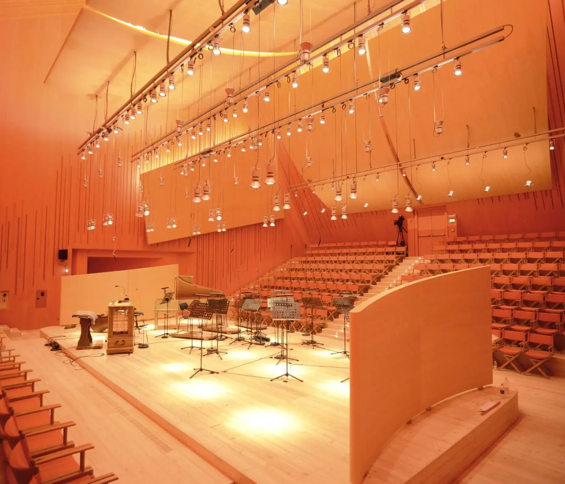 Auditorium in legno con pareti acustiche e gradinate; palco con strumenti musicali, microfoni e spartiti, illuminazione calda arancione.