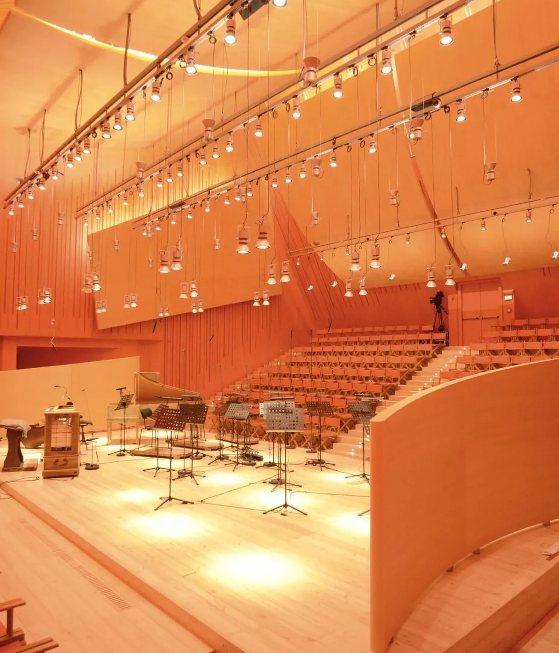 Auditorium in legno con pareti acustiche e gradinate; palco con strumenti musicali, microfoni e spartiti, illuminazione calda arancione.