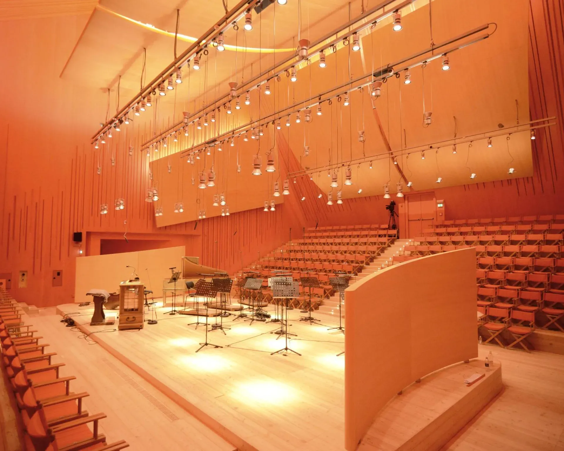 Auditorium in legno con pareti acustiche e gradinate; palco con strumenti musicali, microfoni e spartiti, illuminazione calda arancione.