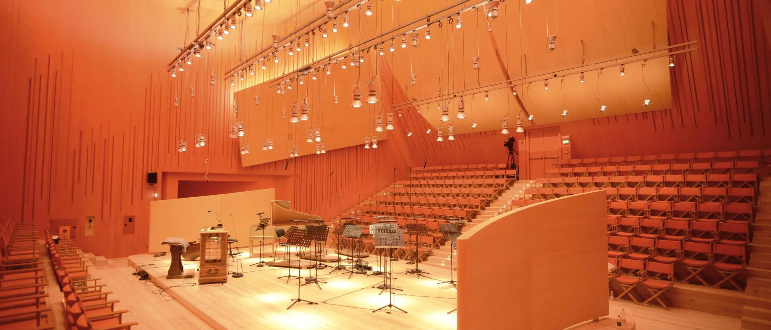 Auditorium in legno con pareti acustiche e gradinate; palco con strumenti musicali, microfoni e spartiti, illuminazione calda arancione.