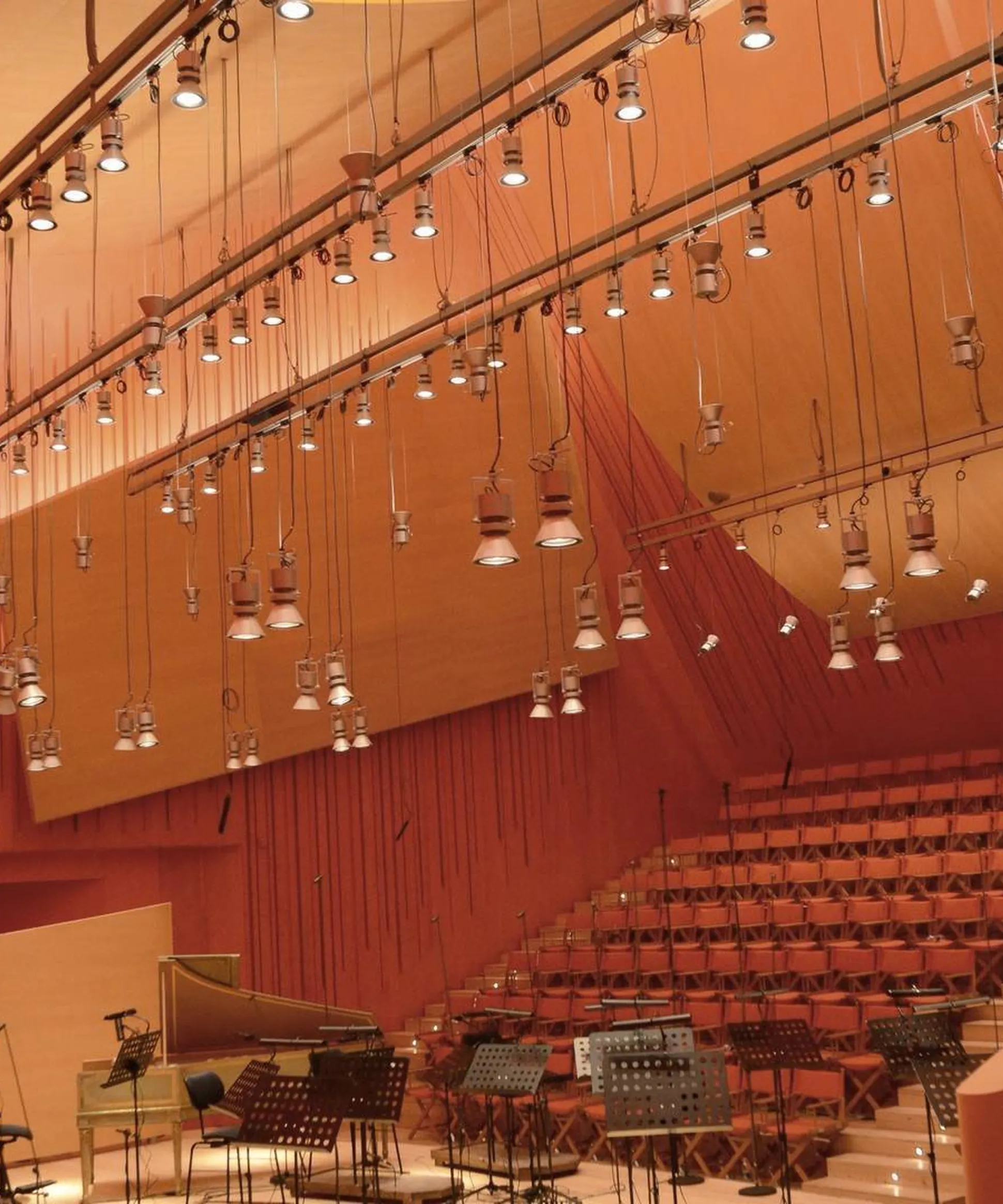 Auditorium in legno con soluzioni acustiche sospese, luci pendenti, palco con cavalletti per spartiti e microfoni, sedute a gradinate.