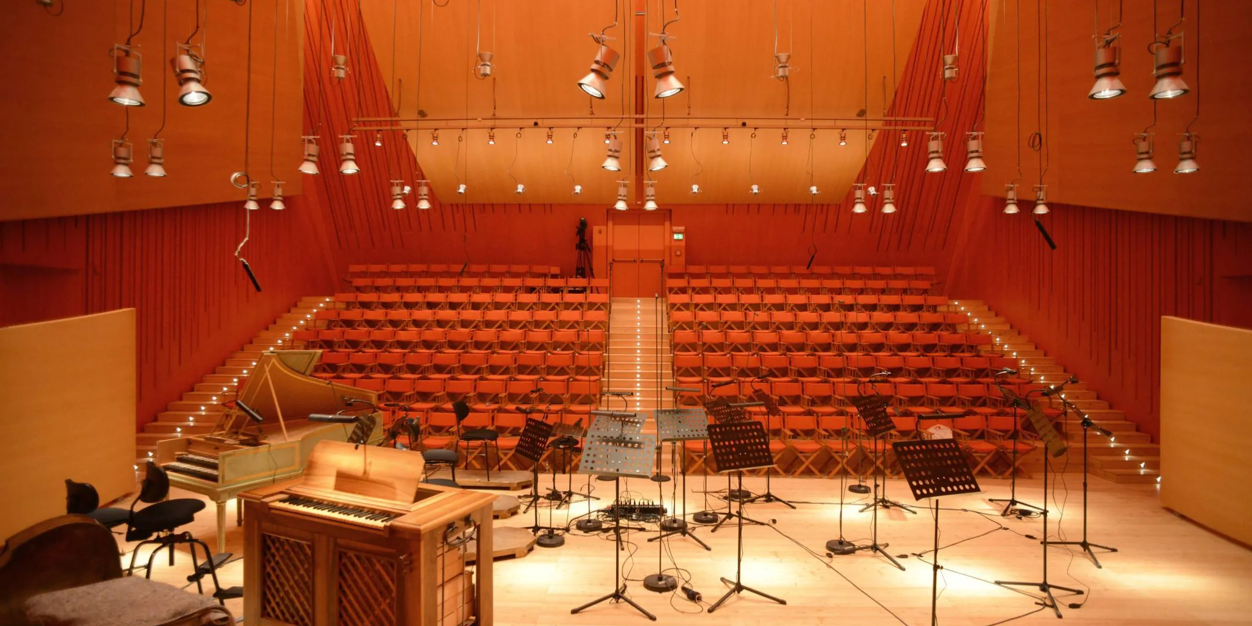 Auditorium in legno con gradinate a rampa, palco attrezzato con pianoforte, microfoni su cavalletti e supporti per spartiti; luci e cavi sospesi dal soffitto.