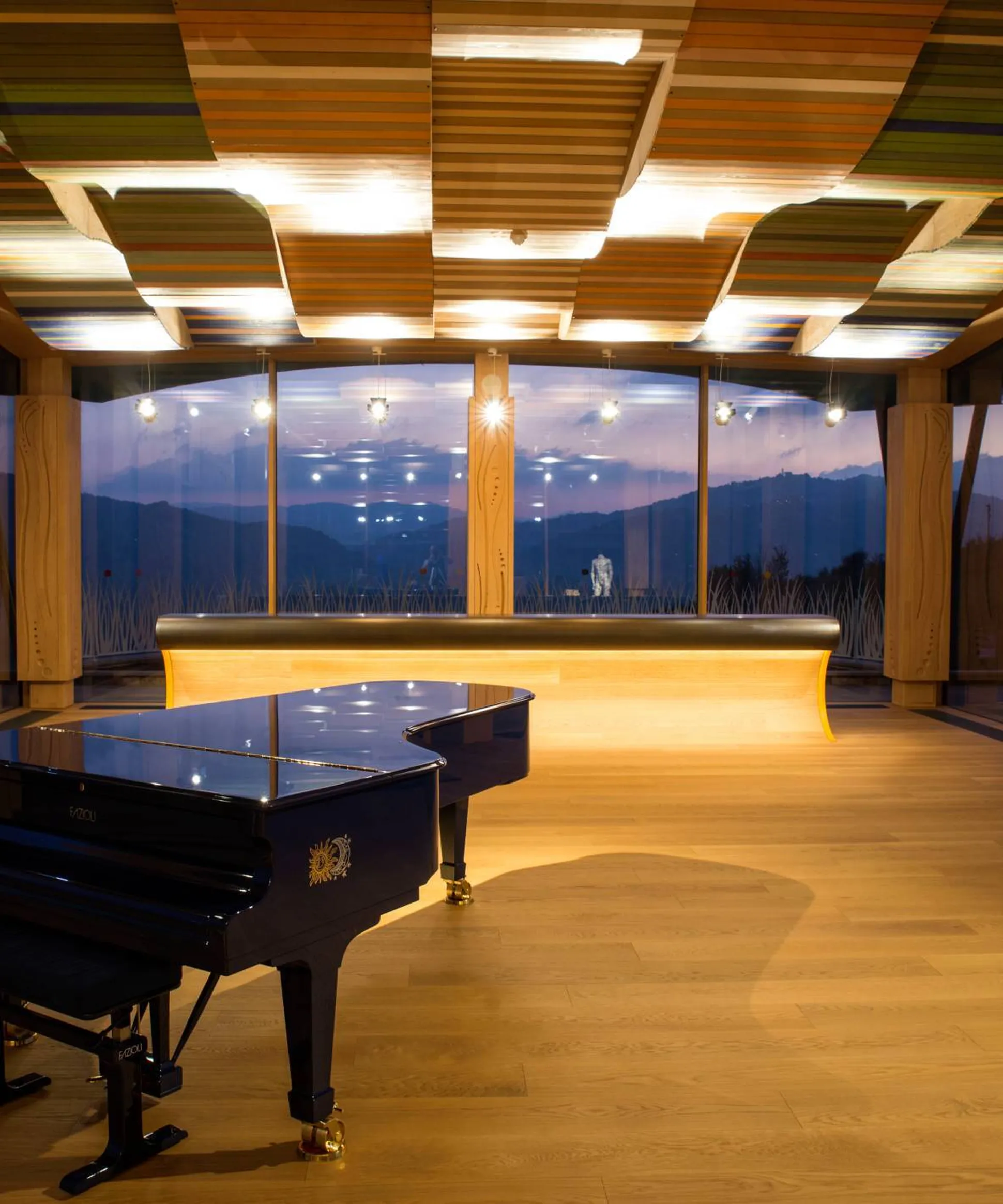 Interno elegante con pianoforte a coda nero al centro, pavimento in legno, pannelli acustici colorati sul soffitto e grandi finestre con vista montane al tramonto.