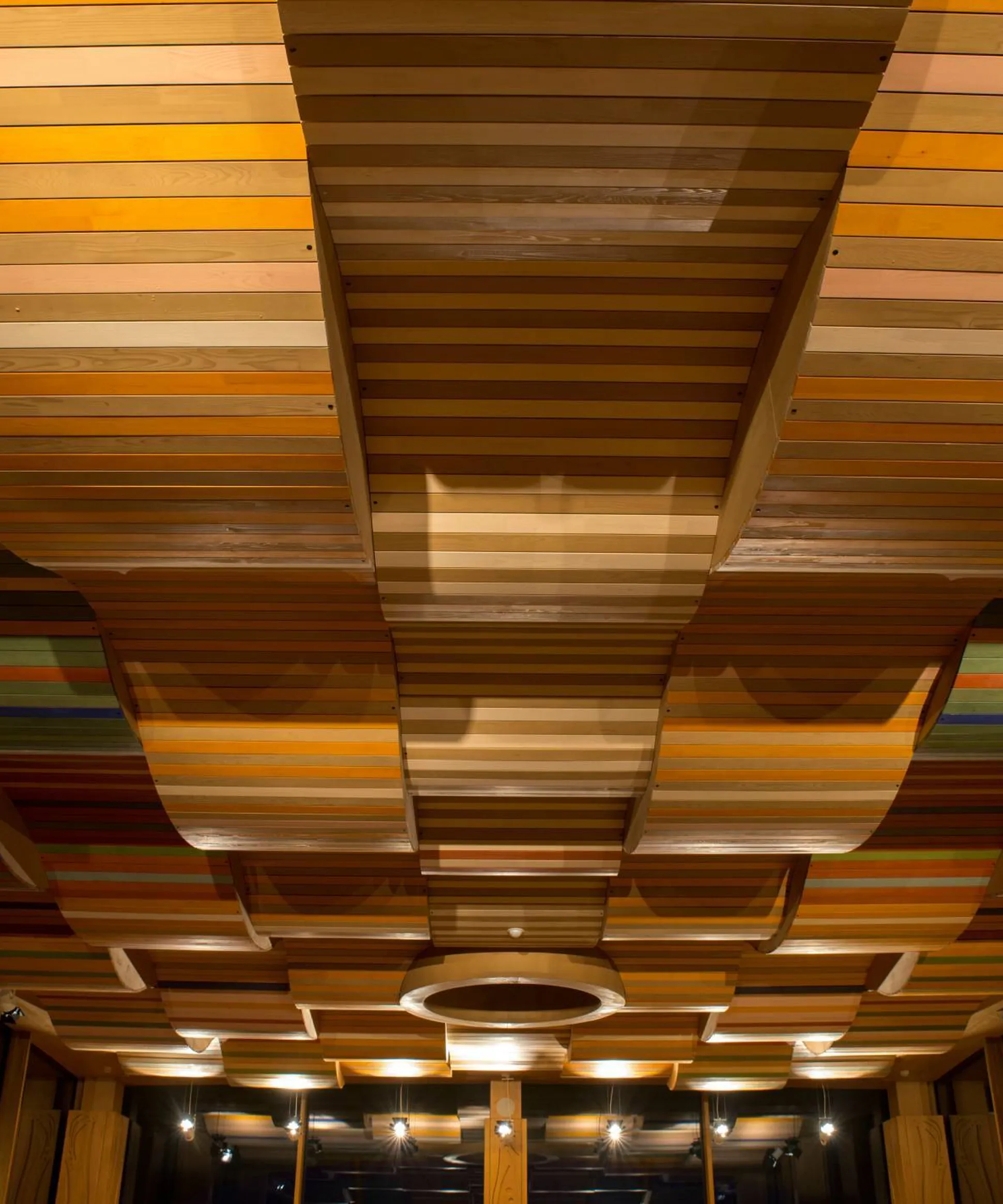 Soffitto di listelli di legno multicolori disposti in pannelli curvi, creando onde. Illuminazione calda enfatizza le texture; ambiente interno con colonne e finestre.