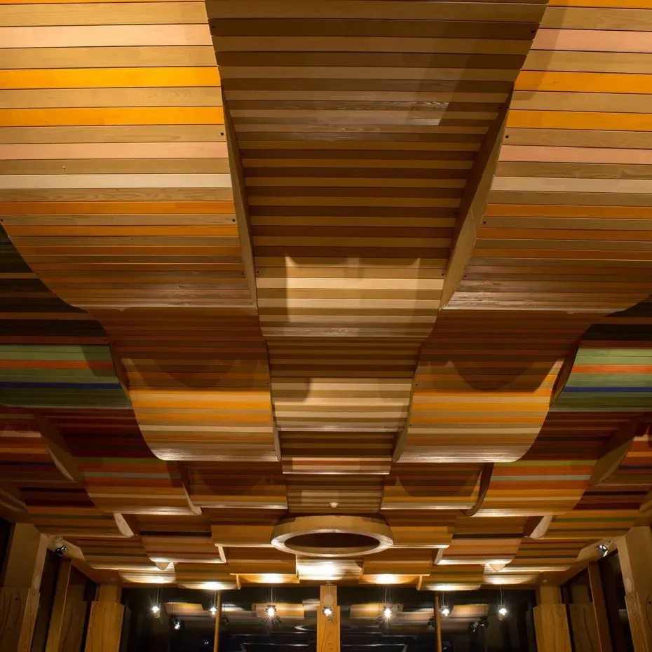 Soffitto di listelli di legno multicolori disposti in pannelli curvi, creando onde. Illuminazione calda enfatizza le texture; ambiente interno con colonne e finestre.