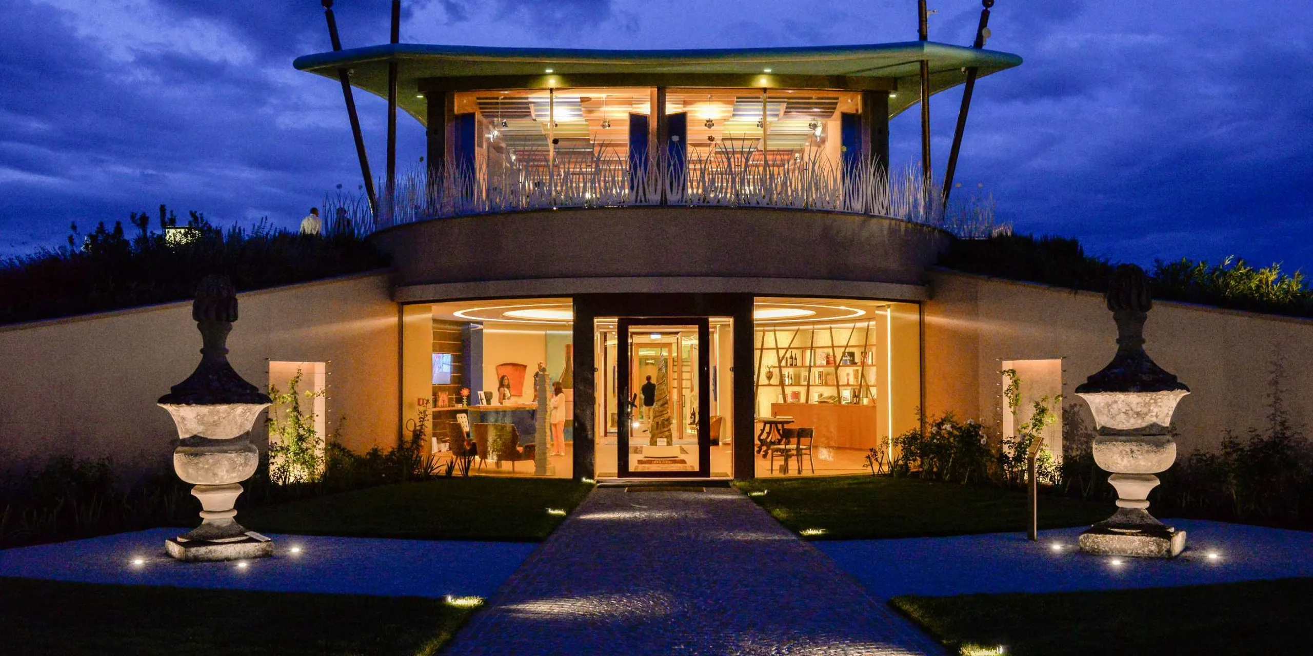 Edificio moderno con ingresso in vetro e sala interna illuminata; terrazza ovale sul tetto, percorso lastricato con luci e due urne decorative ai lati.