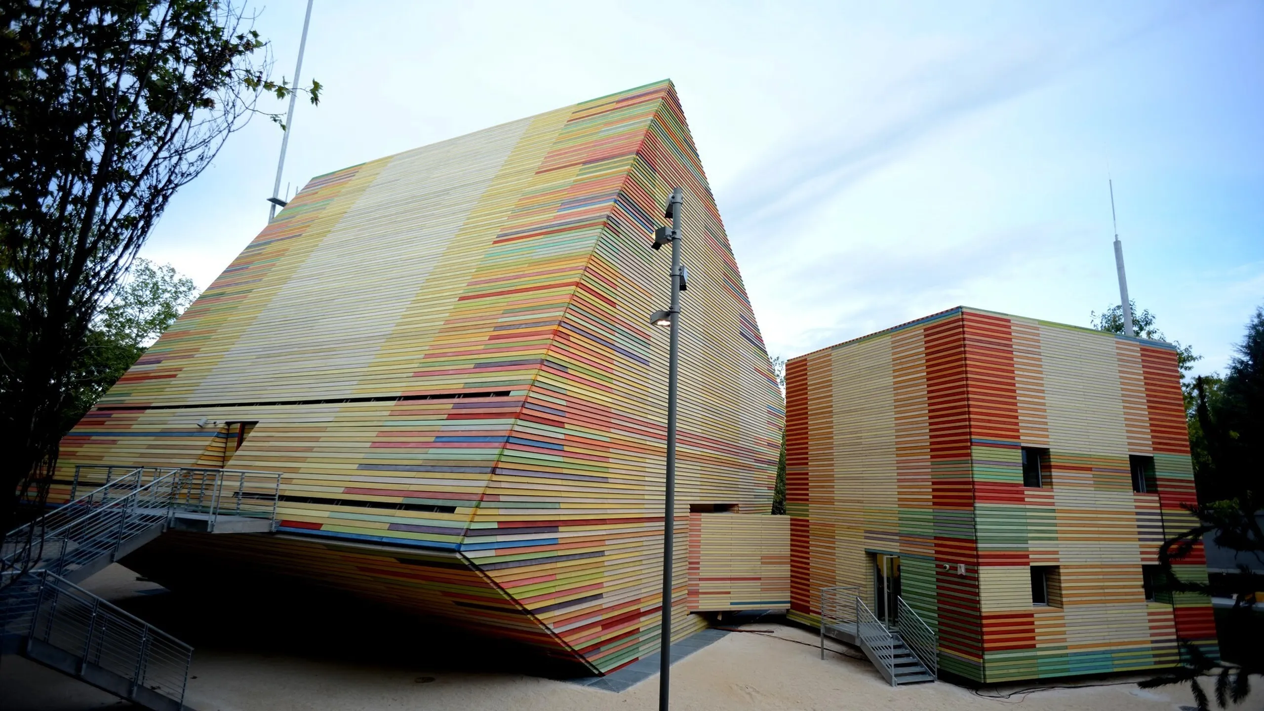 Edificio moderno rivestito di listelli orizzontali multicolori, con volume inclinato e volume rettangolare, accessi pedonali in metallo.