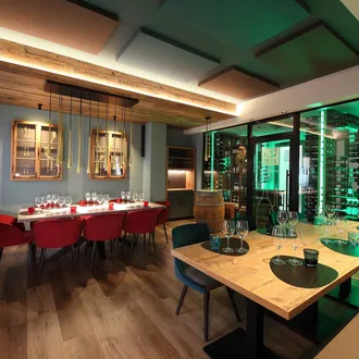 Interno Silenzia: sala ristorante con pannelli acustici sul soffitto, tavolo lungo bianco con sedie rosse e cantina illuminata di verde dietro vetri.
