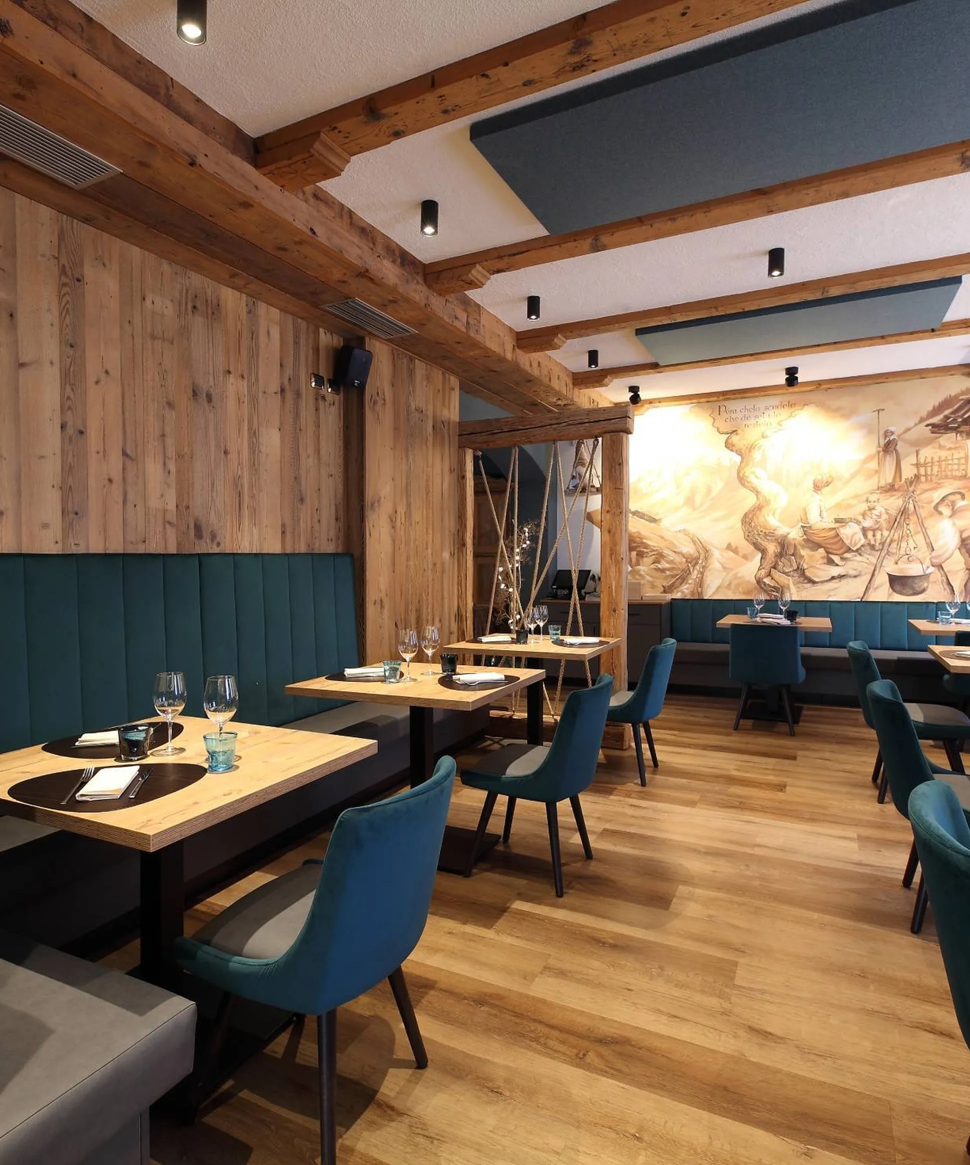 Ristorante con travi in legno, panche imbottite e sedie blu, tavoli in legno e un grande murale rustico sullo sfondo.