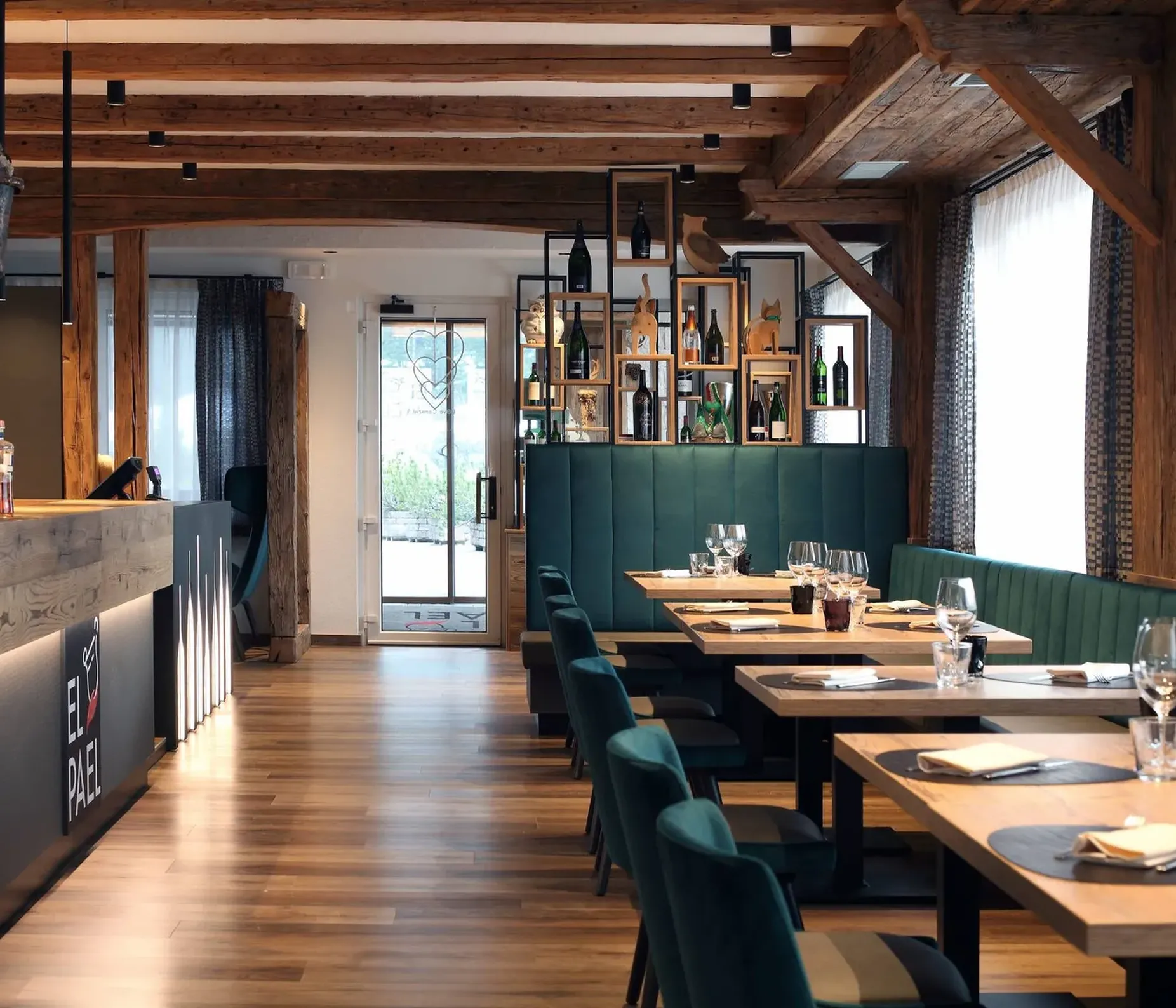 Interno di ristorante con travi in legno, bancone bar a sinistra, scaffali con bottiglie alle spalle, divani verdi e tavoli di legno con bicchieri.