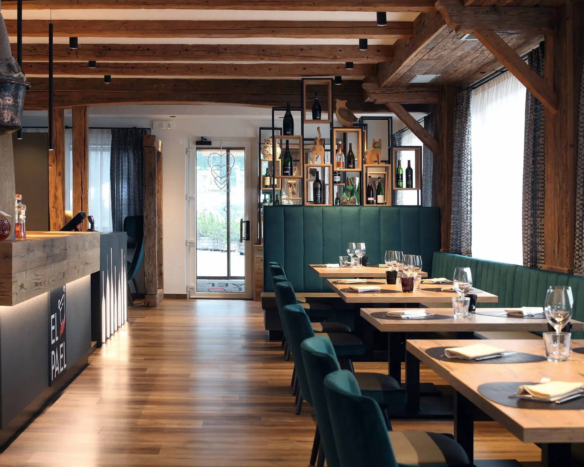 Interno di ristorante con travi in legno, bancone bar a sinistra, scaffali con bottiglie alle spalle, divani verdi e tavoli di legno con bicchieri.