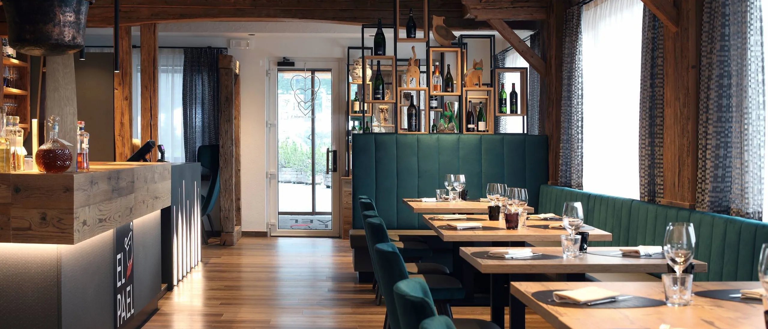 Interno di ristorante con travi in legno, bancone bar a sinistra, scaffali con bottiglie alle spalle, divani verdi e tavoli di legno con bicchieri.