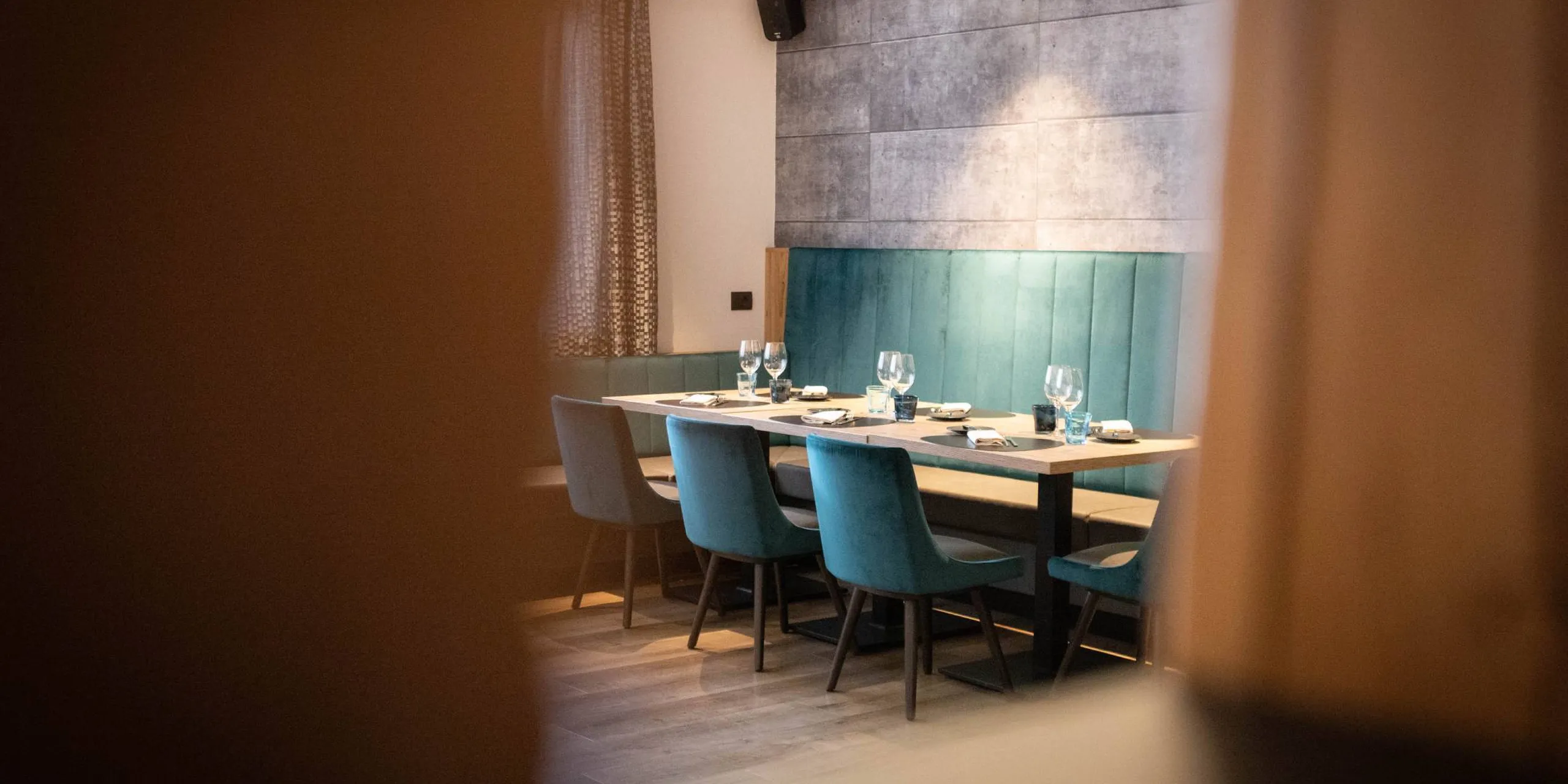 Sala ristorante moderna con tavolo lungo apparecchiato: bicchieri e piatti, sedie blu-teal, panca verde acqua e pareti di pannelli grigi fonoassorbenti; sfocato.