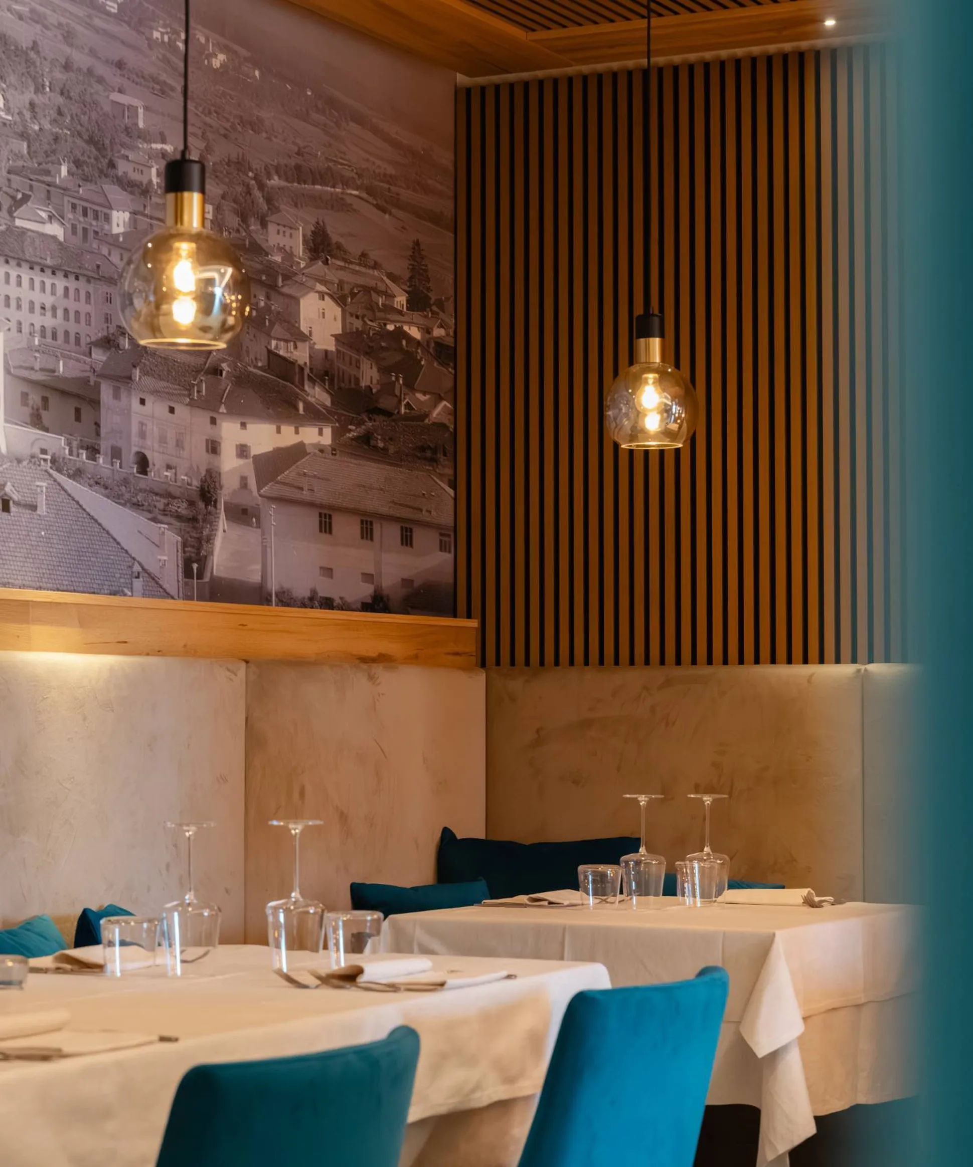 Interni moderni di ristorante: tavoli con tovaglie bianche, sedie azzurre, lampade a bulbo, pareti in legno a strisce e murale urbano in bianco e nero.