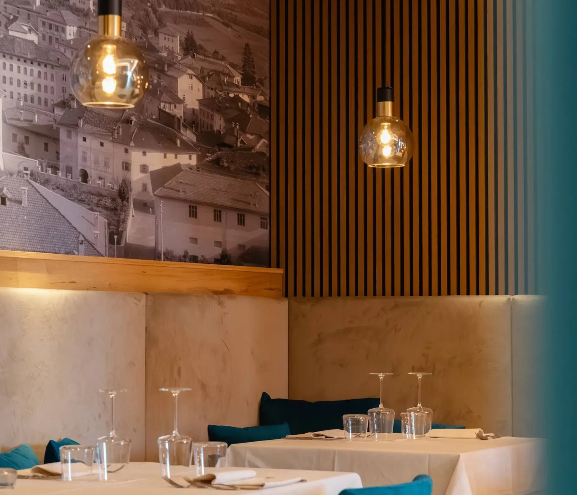 Interni moderni di ristorante: tavoli con tovaglie bianche, sedie azzurre, lampade a bulbo, pareti in legno a strisce e murale urbano in bianco e nero.