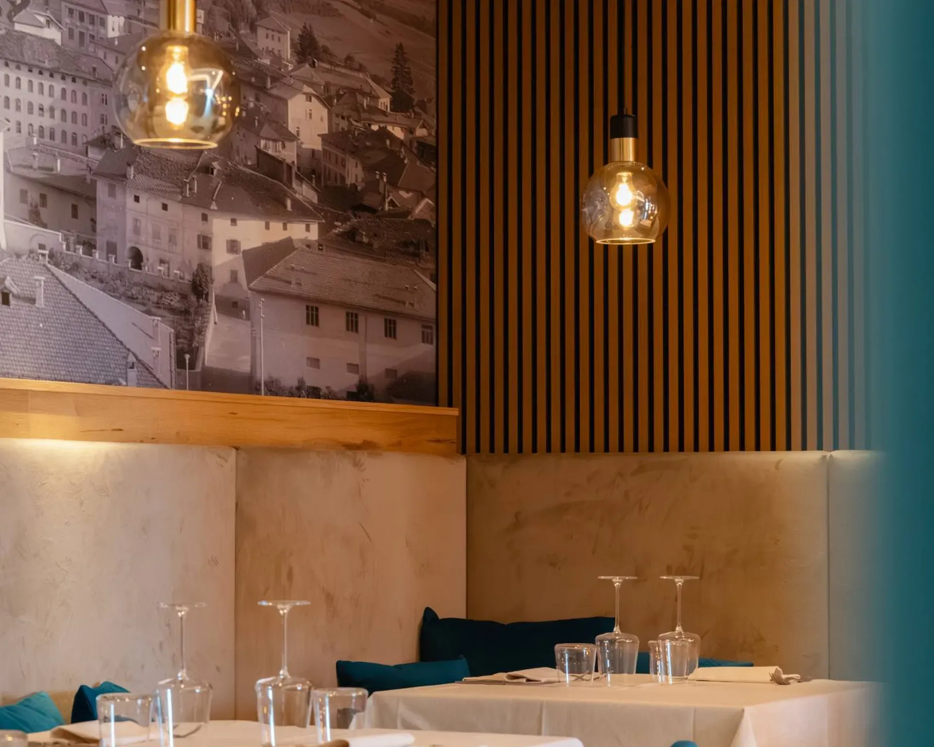 Interni moderni di ristorante: tavoli con tovaglie bianche, sedie azzurre, lampade a bulbo, pareti in legno a strisce e murale urbano in bianco e nero.