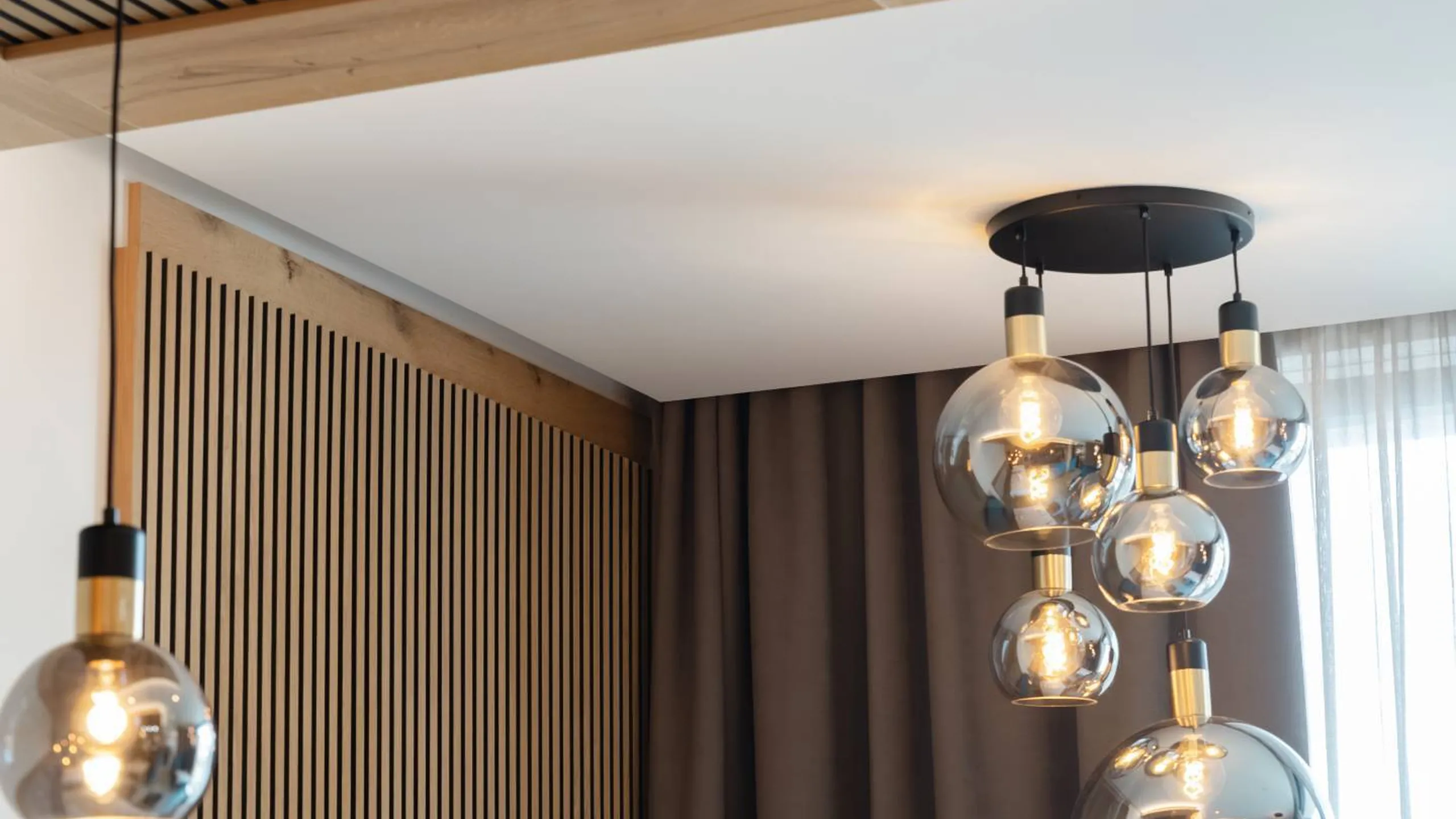 Interno moderno con pannelli fonoassorbenti a listelli in legno, soffitto con travi e lampade pendenti a sfere di vetro. Tende grigie e divano blu.