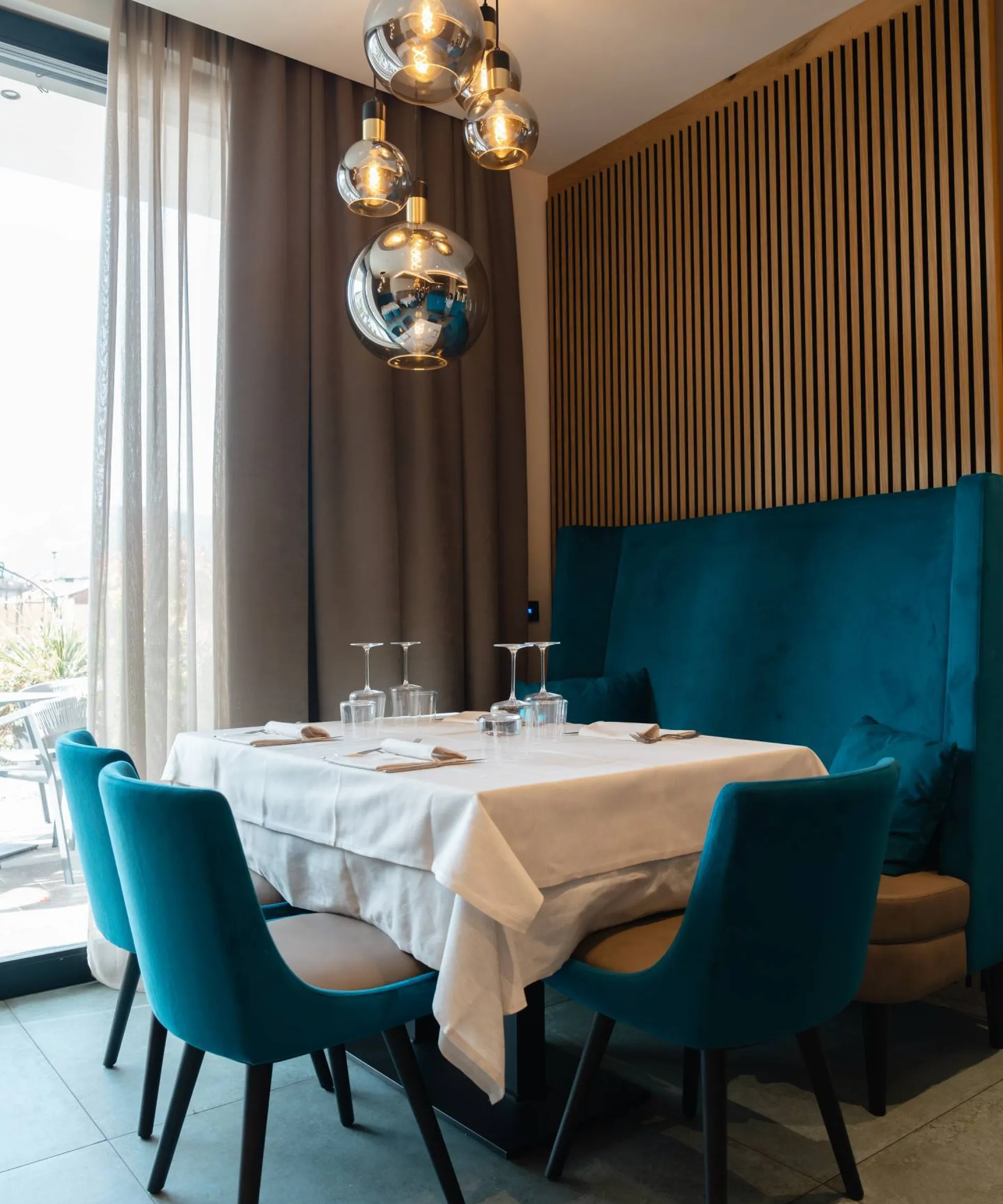 Sedie imbottite turchesi attorno a un tavolo bianco con tovaglia, in un ristorante moderno, lampade pendenti a sfere e parete in listelli di legno.