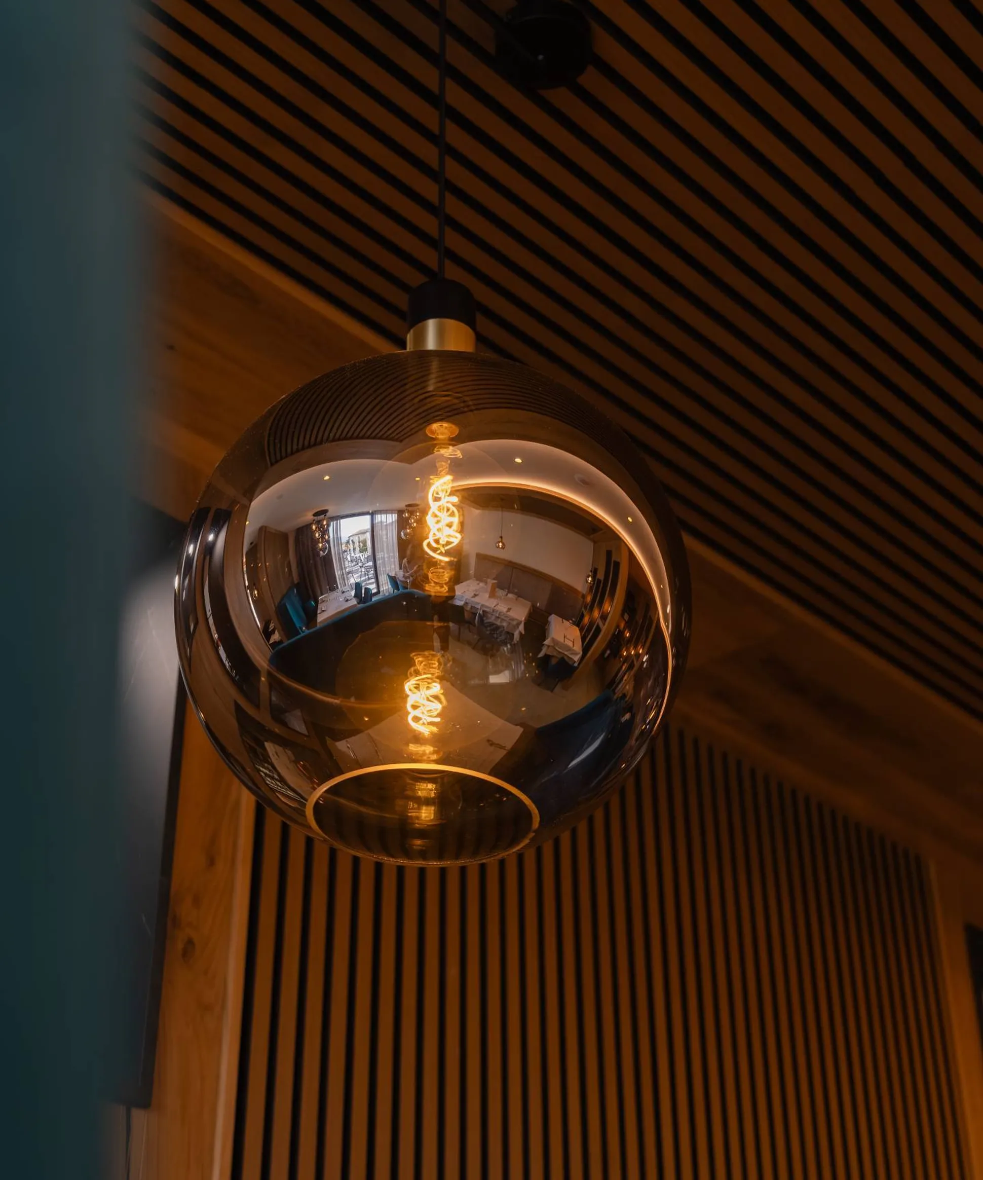 Lampadario a sfera in vetro riflettente appeso al soffitto; riflette l’interno con tavoli, sedie e pareti di listelli di legno.