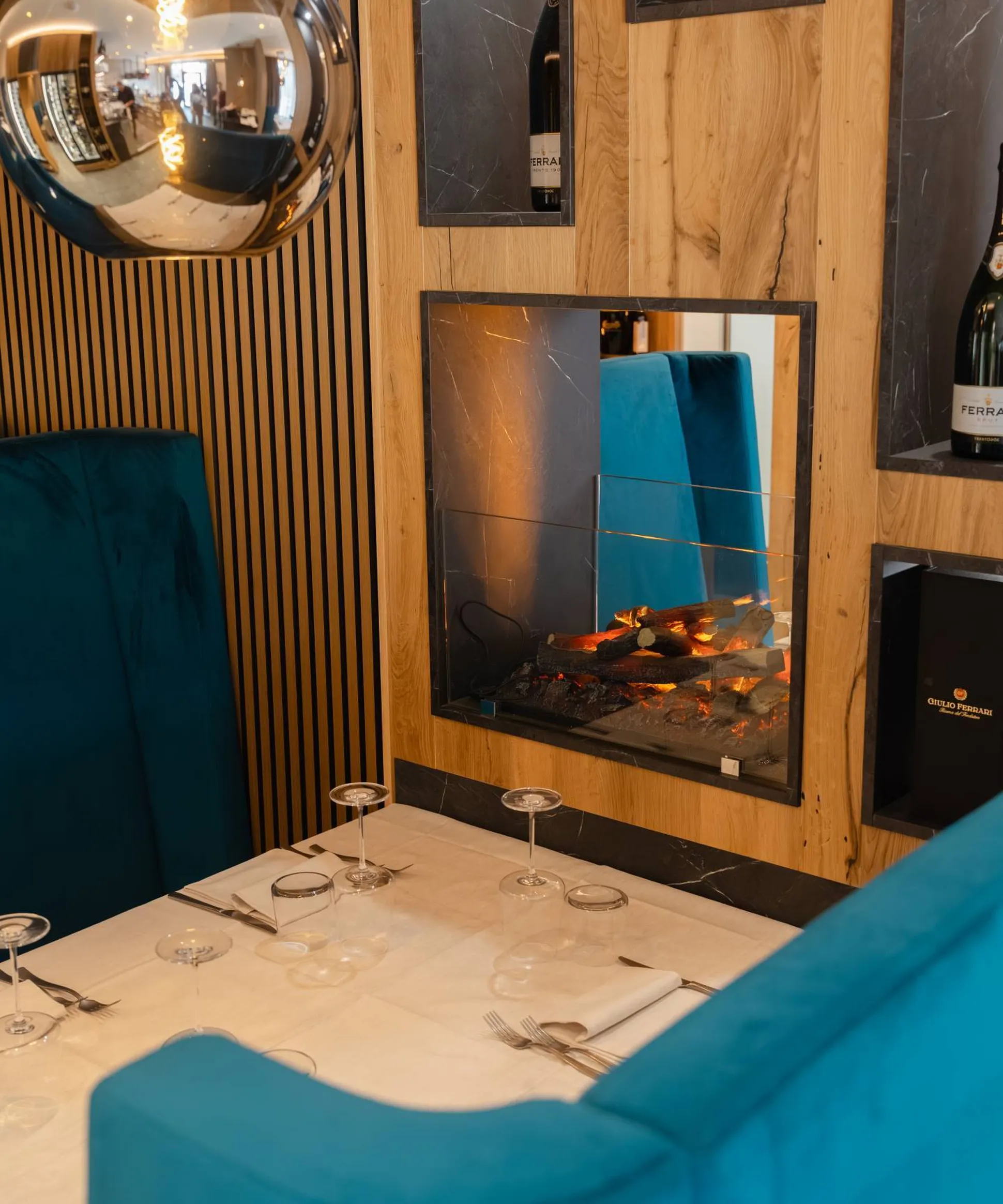 Zona pranzo con tavolo bianco, posate e bicchieri, divani blu, camino moderno incastonato in pannelli di legno; lampada sferica riflette nell'ambiente.