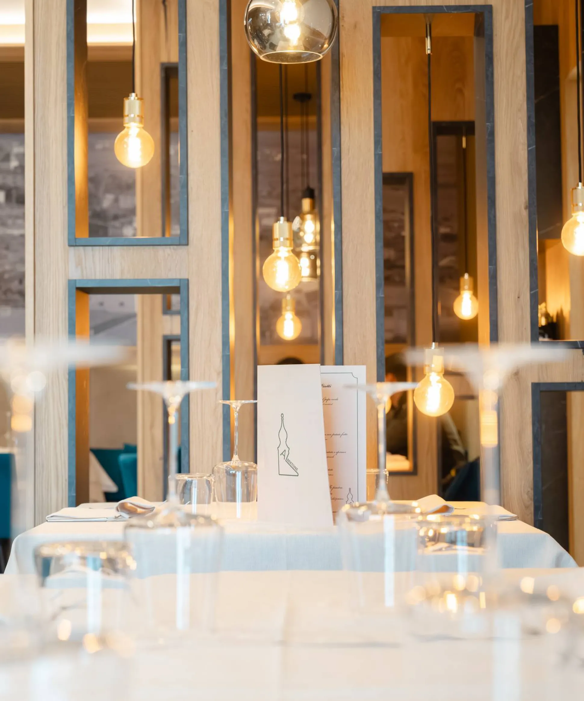 Interno di ristorante moderno con pannelli in legno e cornici, luci a bulbo pendenti, tavolo apparecchiato con bicchieri e menù.