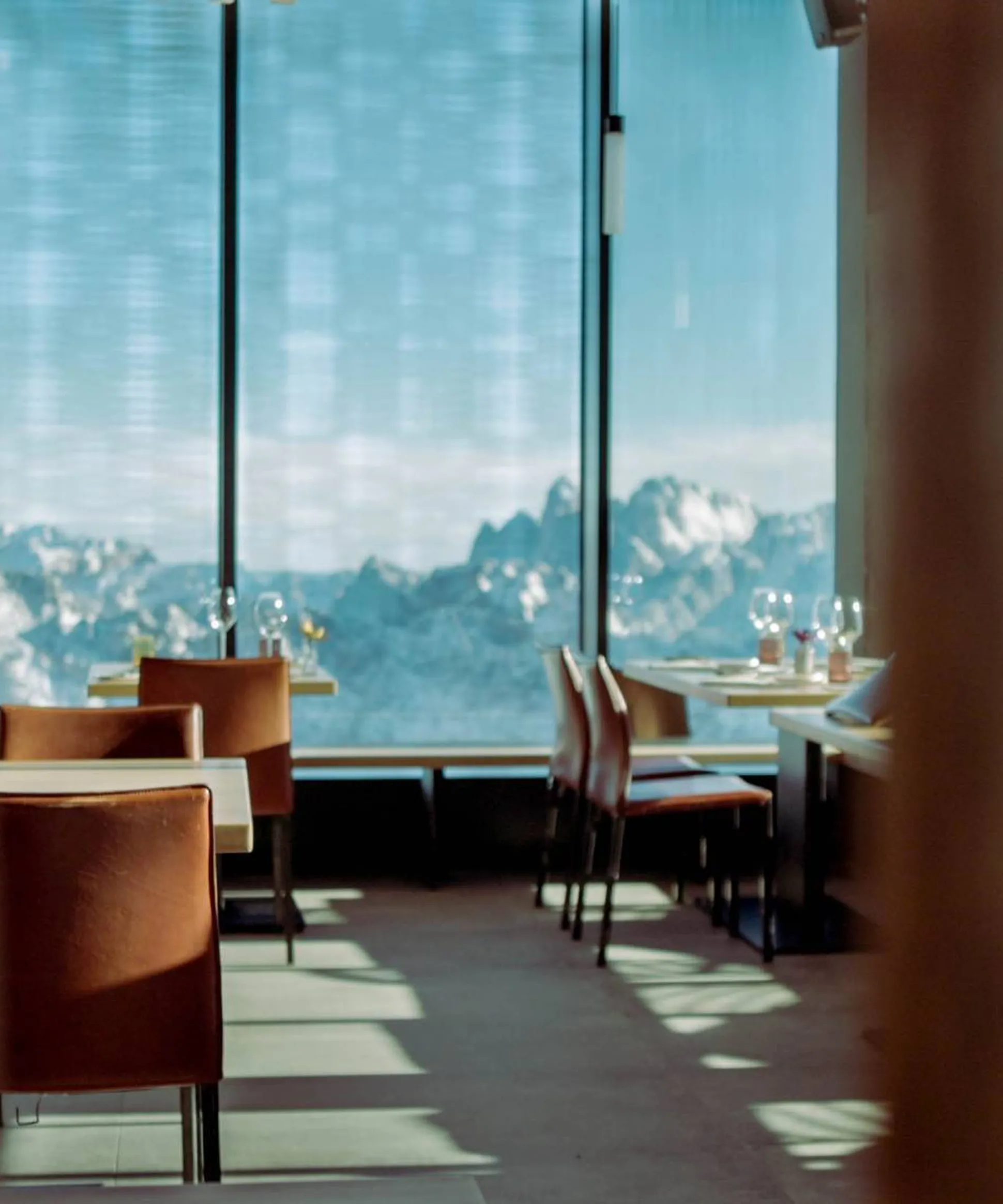Ristorante con tavoli apparecchiati e sedie arancioni, vista su montagne innevate dalle grandi vetrate, luce naturale e ombre sul pavimento.