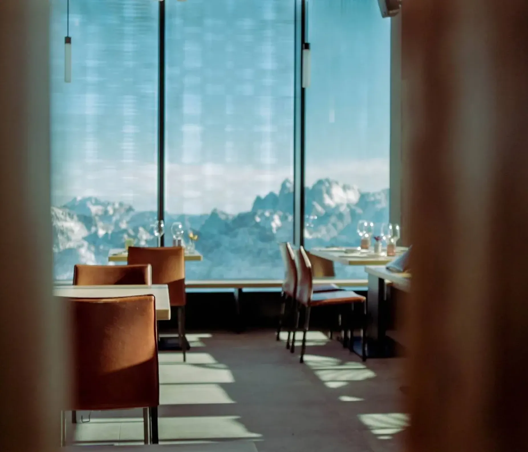 Ristorante con tavoli apparecchiati e sedie arancioni, vista su montagne innevate dalle grandi vetrate, luce naturale e ombre sul pavimento.