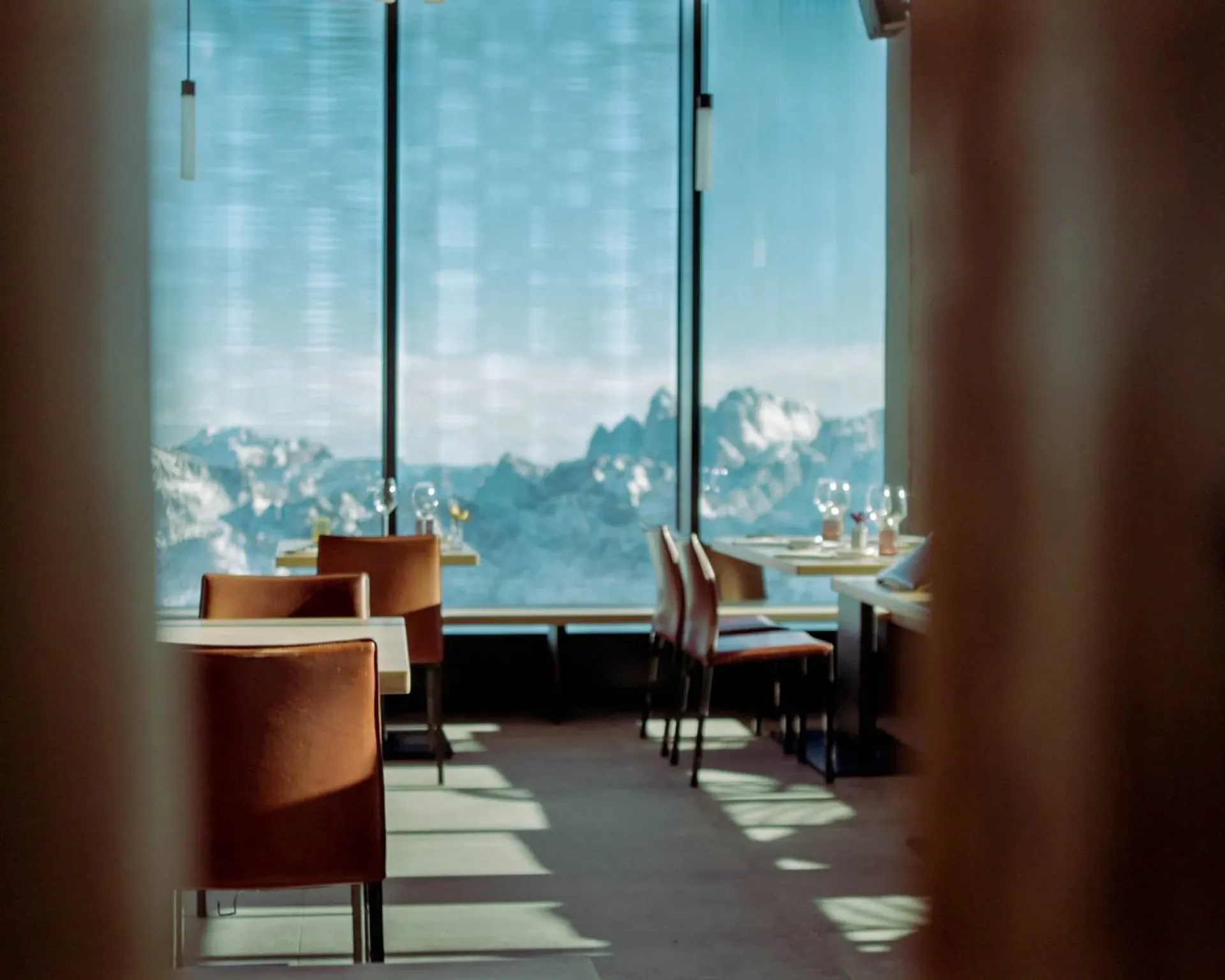 Ristorante con tavoli apparecchiati e sedie arancioni, vista su montagne innevate dalle grandi vetrate, luce naturale e ombre sul pavimento.