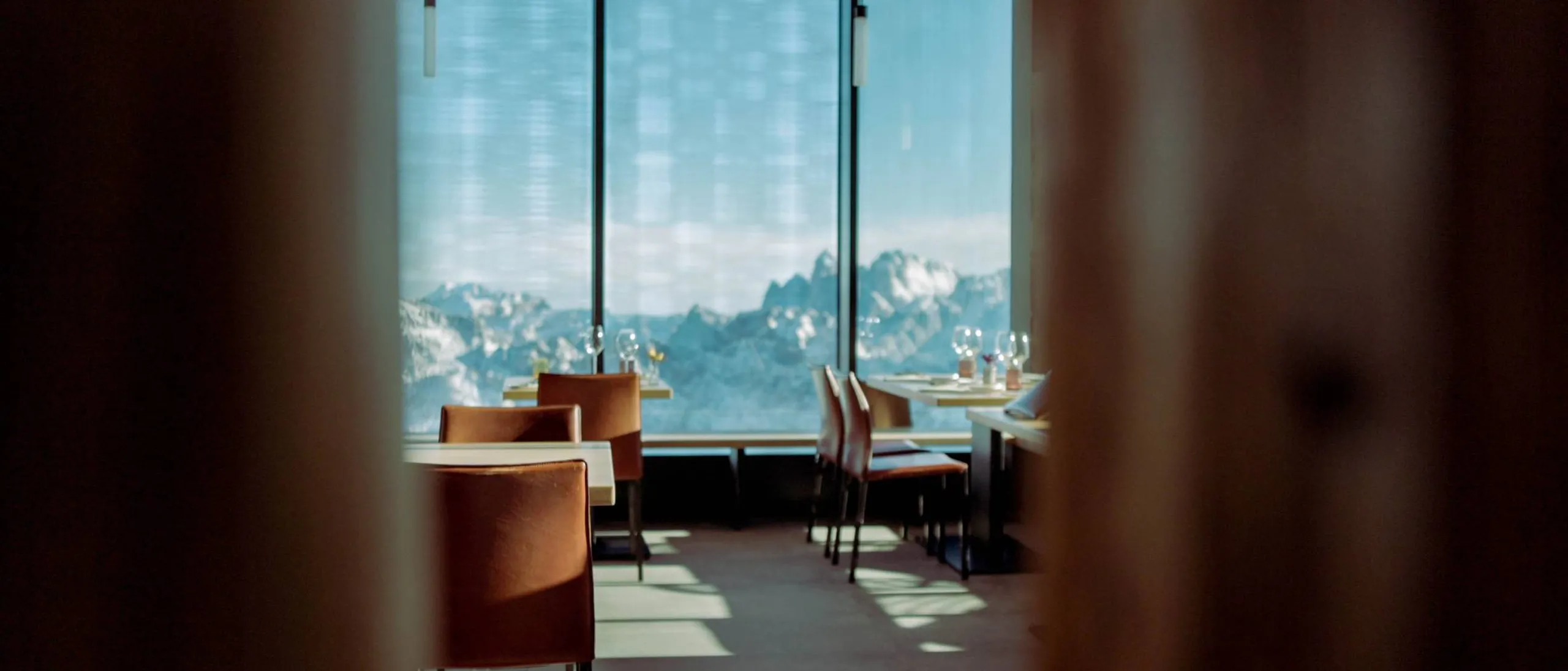 Ristorante con tavoli apparecchiati e sedie arancioni, vista su montagne innevate dalle grandi vetrate, luce naturale e ombre sul pavimento.