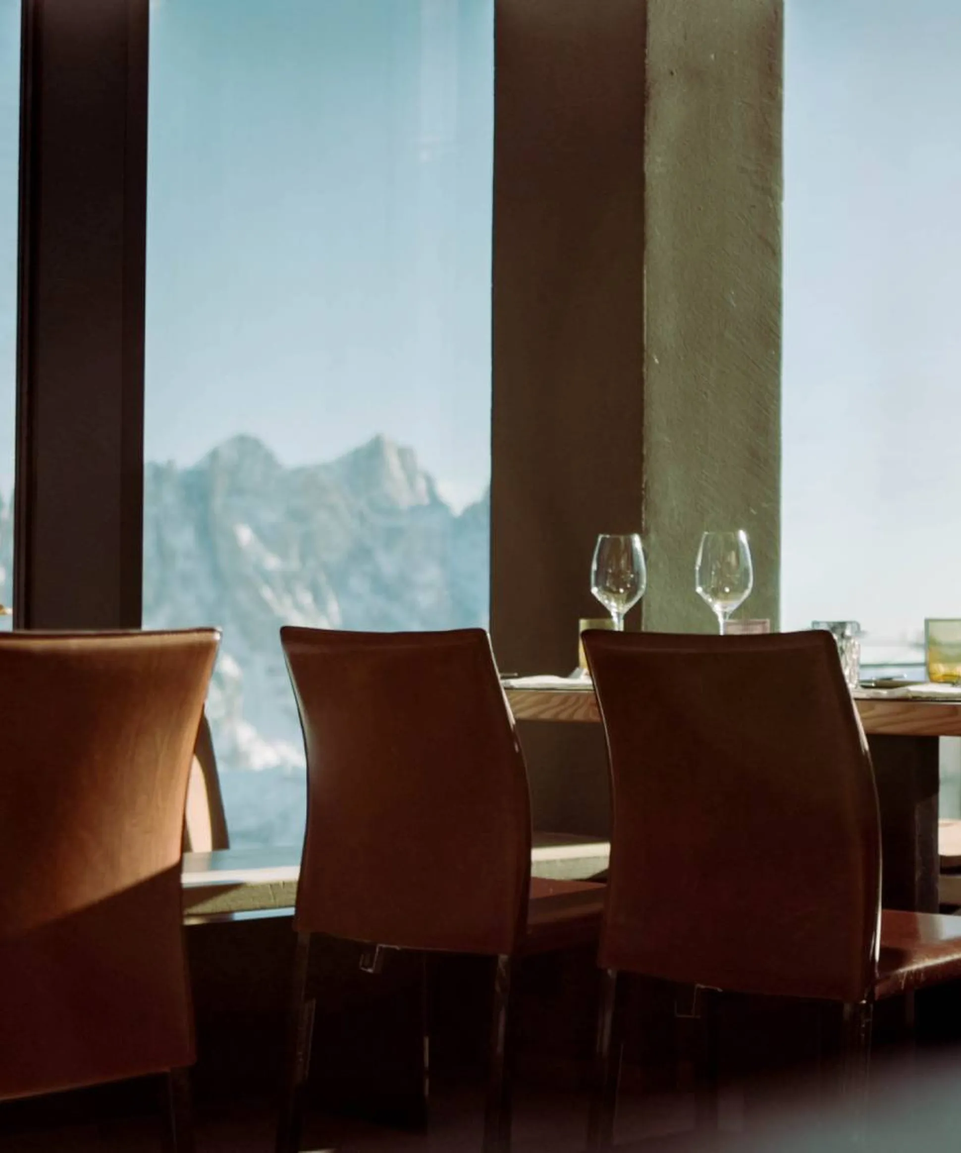 Ristorante moderno: tavoli apparecchiati, sedie marrone in pelle, grandi finestre con vista sulle montagne e bicchieri sul tavolo.