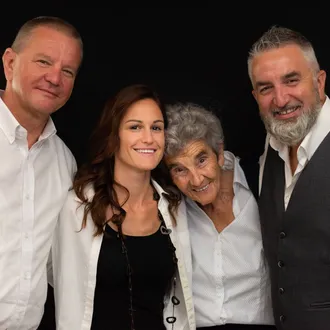 Quattro individui sorridenti in posa contro uno sfondo nero: due uomini in camicia bianca, una giovane donna con blazer bianco e top nero, e una donna anziana.