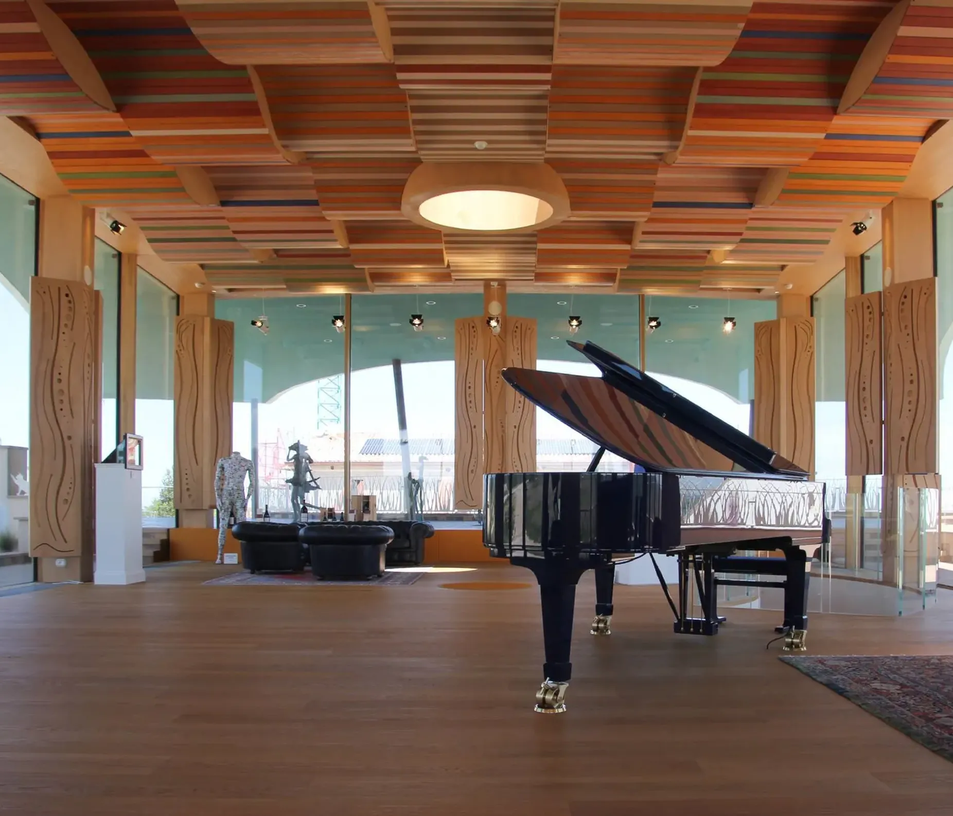 Interno moderno con pianoforte a coda nero, coperchio aperto, soffitto a listelli colorati, colonne in legno e grandi vetrate; divano rosso in zona lounge.