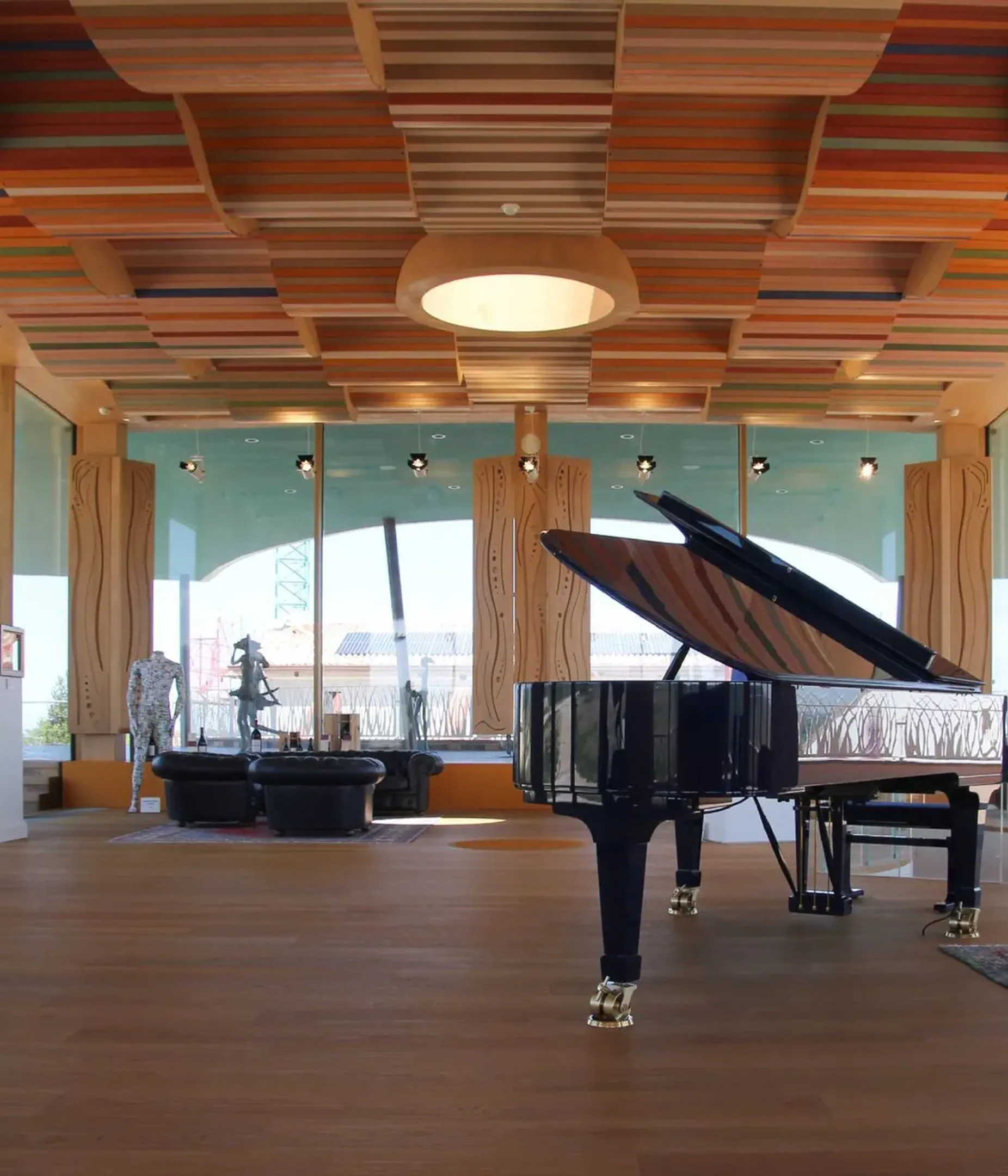 Interno moderno con pianoforte a coda nero, coperchio aperto, soffitto a listelli colorati, colonne in legno e grandi vetrate; divano rosso in zona lounge.